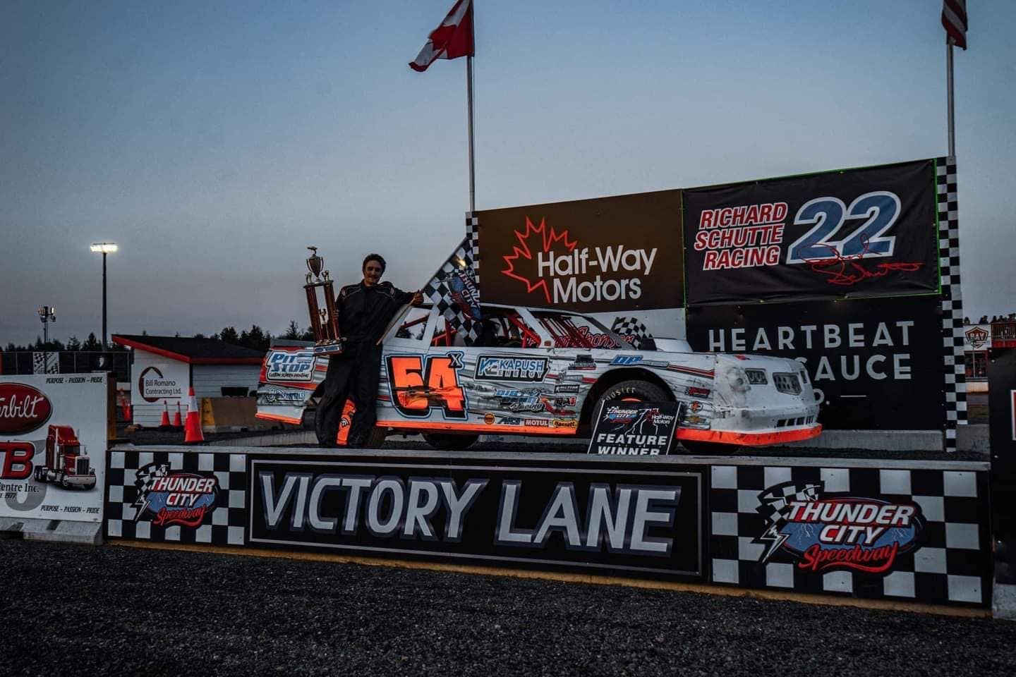 Race car driver celebrating a win on the victory lane podium, holding a trophy. The car is white with orange accents, at Thunder City Speedway.