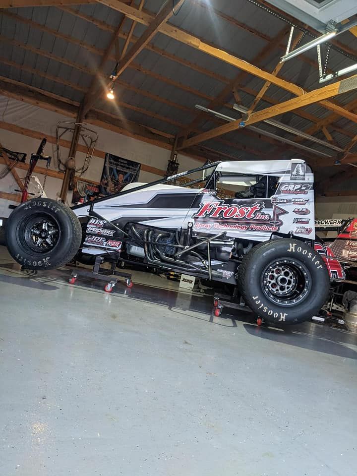 Race car inside a garage. The car is gray, white, and black, with large black tires and sponsor decals.