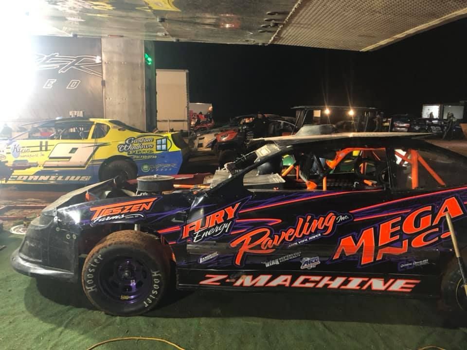 Two race cars, one black and one yellow, in a dimly lit racetrack garage. The black car has purple accents and sponsor logos.