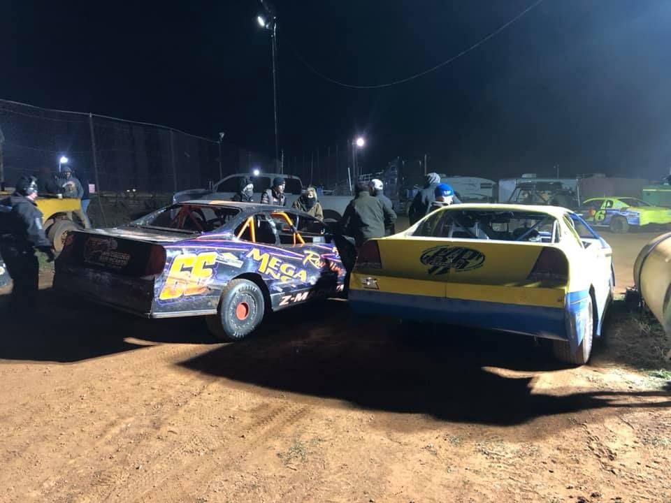 Two stock cars side by side at a dirt track at night. People gather around the cars. The car on the left is purple, the one on the right is yellow.