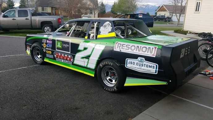Black and green race car with the number 27 parked on a driveway. The car has sponsor logos and is set against a suburb.