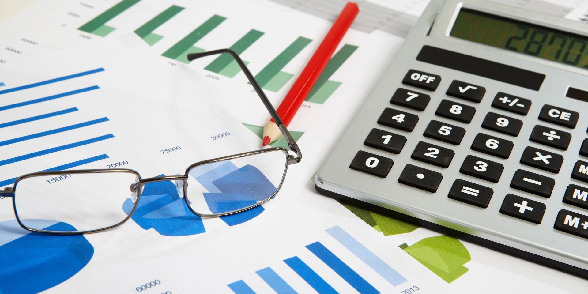 Calculator, eyeglasses, and a pencil on top of financial reports with bar graphs.