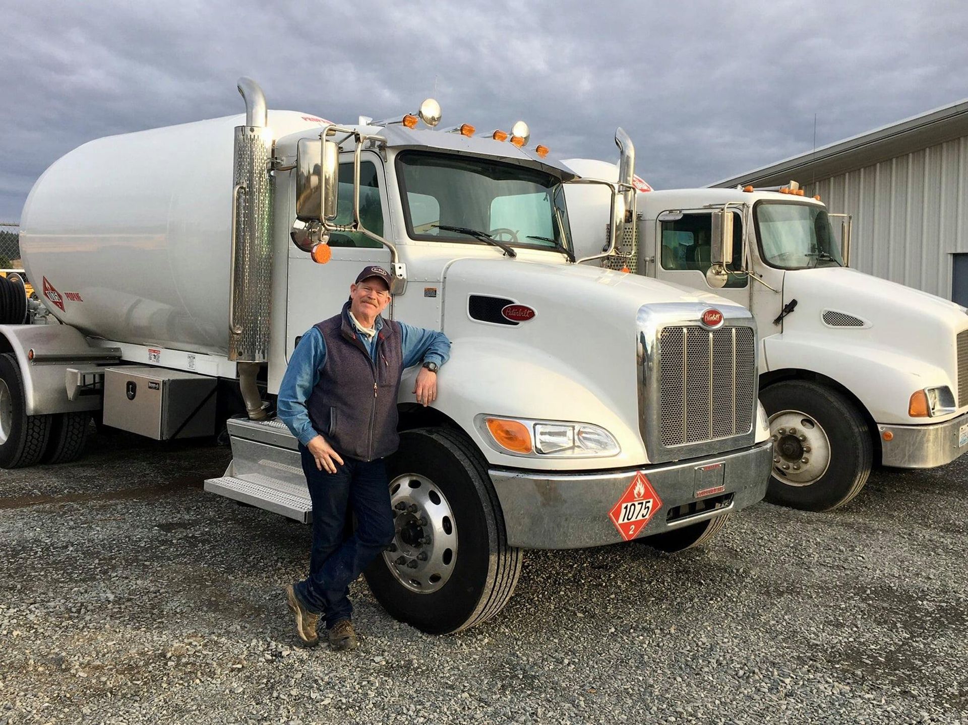 Woman stands by white tanker truck, smiles. Another truck in the background. Gravel lot, cloudy sky.