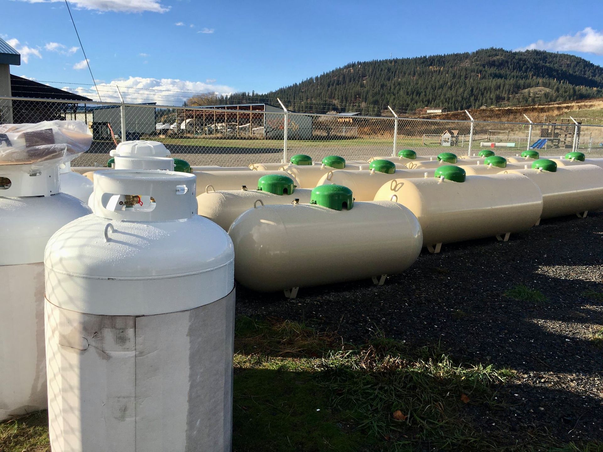 Propane tanks lined up outdoors, tan and white, under a blue sky, with a mountain in the background.