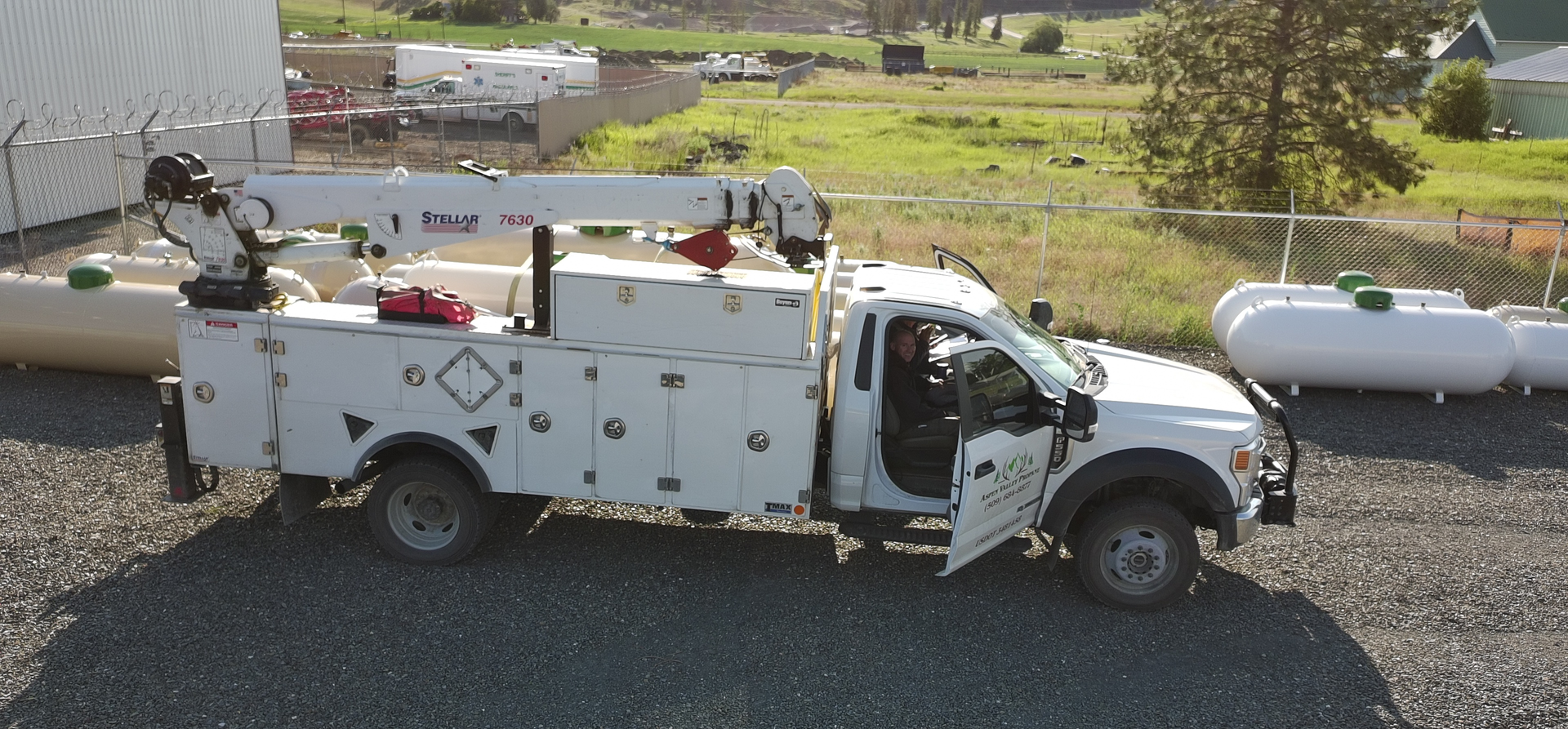 White utility truck with crane parked on gravel, door open. Green grassy field and tanks in the background.