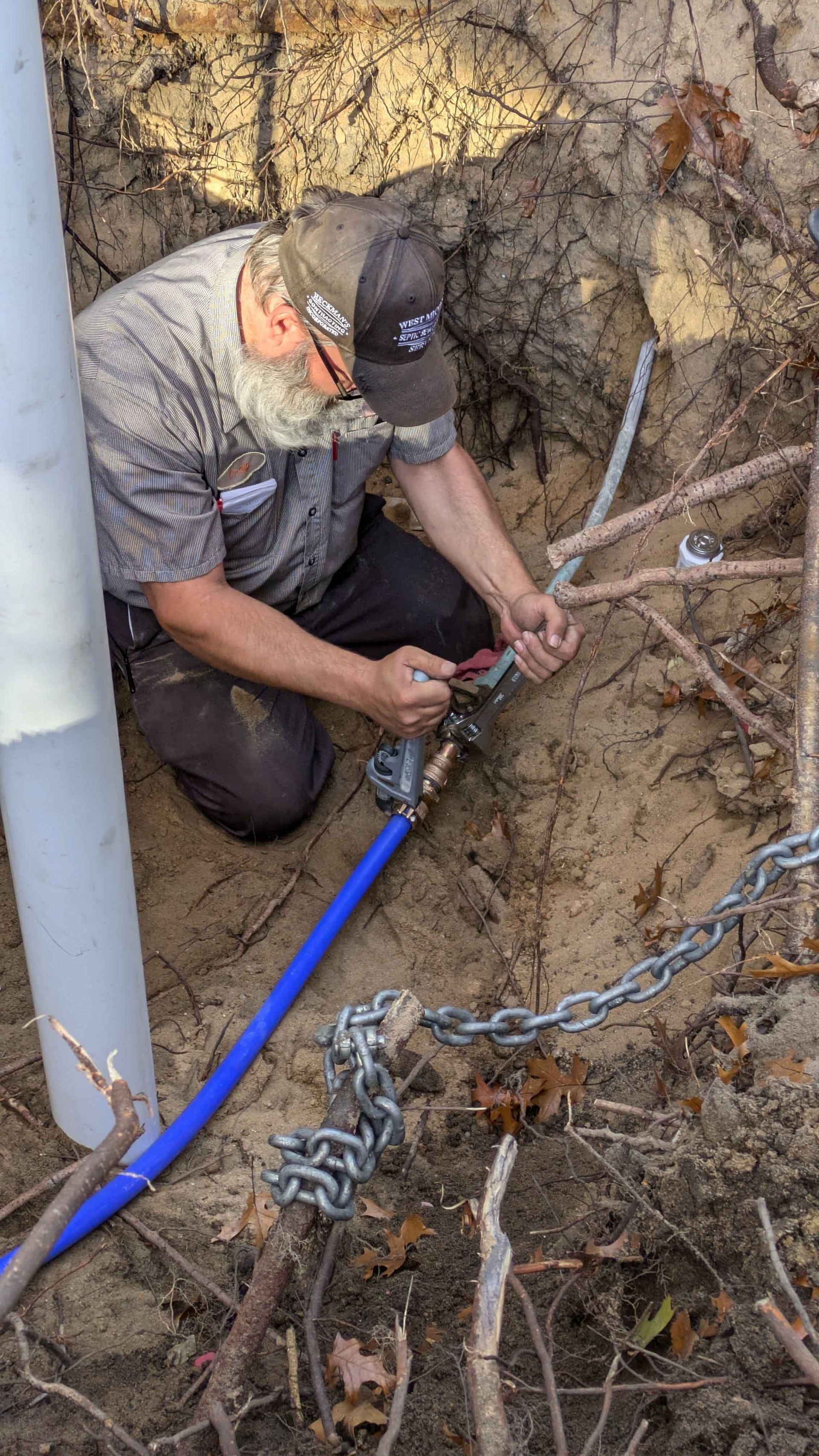 Man in camo working on pipe in a dirt hole outdoors, with a white pole, blue hose, and chains.