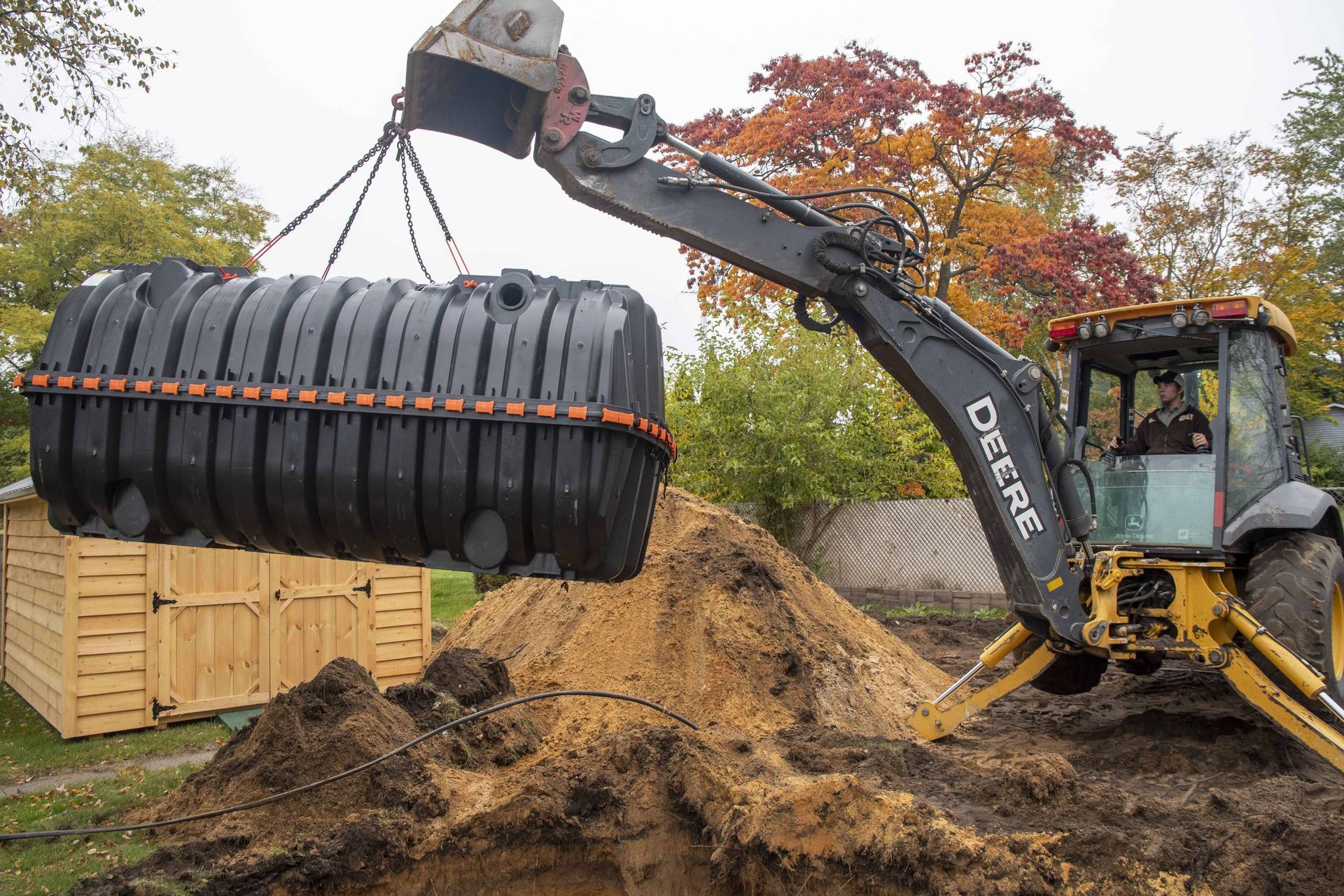 John Deere backhoe lifting a black septic tank over a dirt pit in a yard.