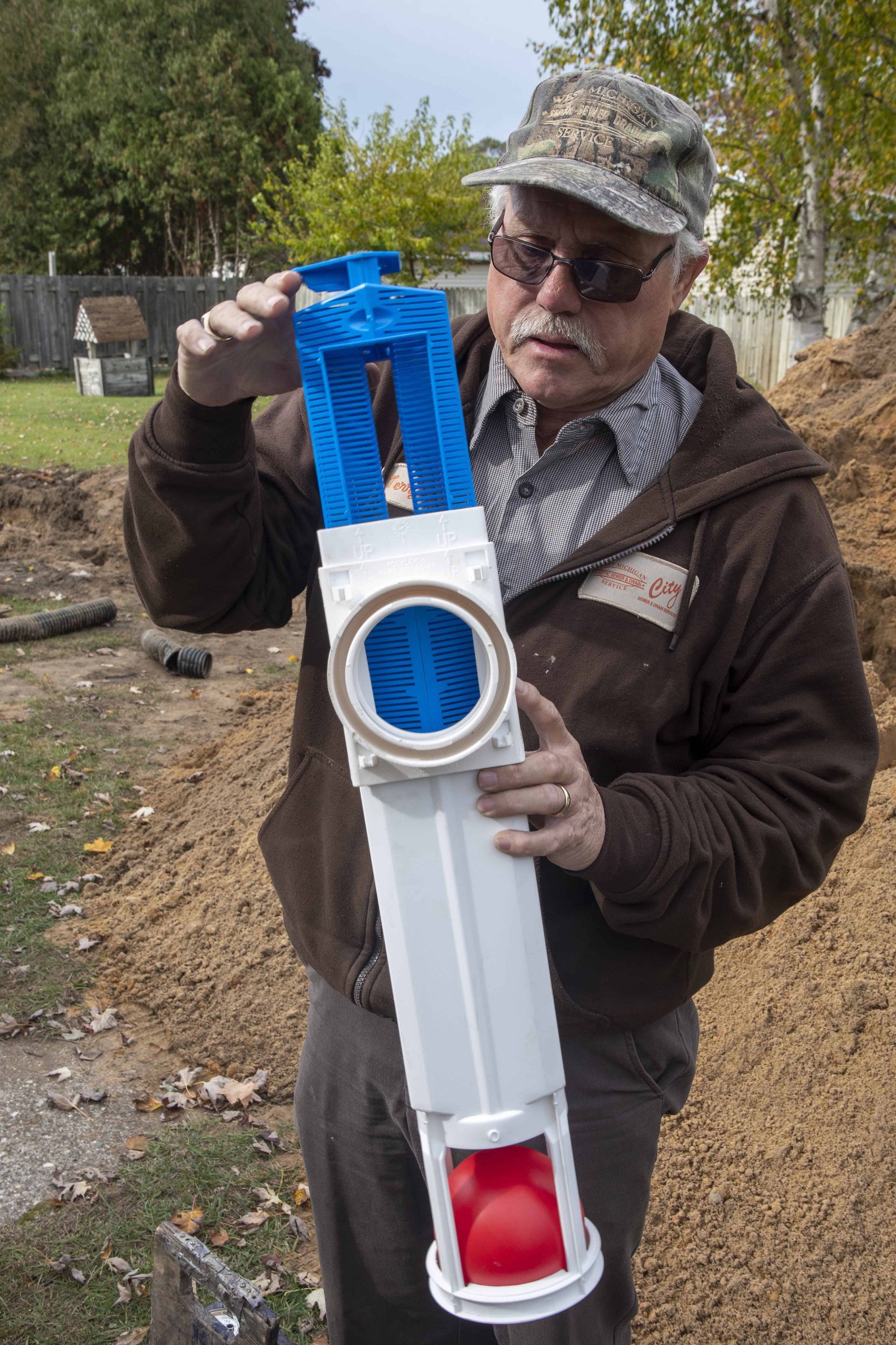Man holding a blue and white drainage device. He wears a brown jacket and hat, outdoors.