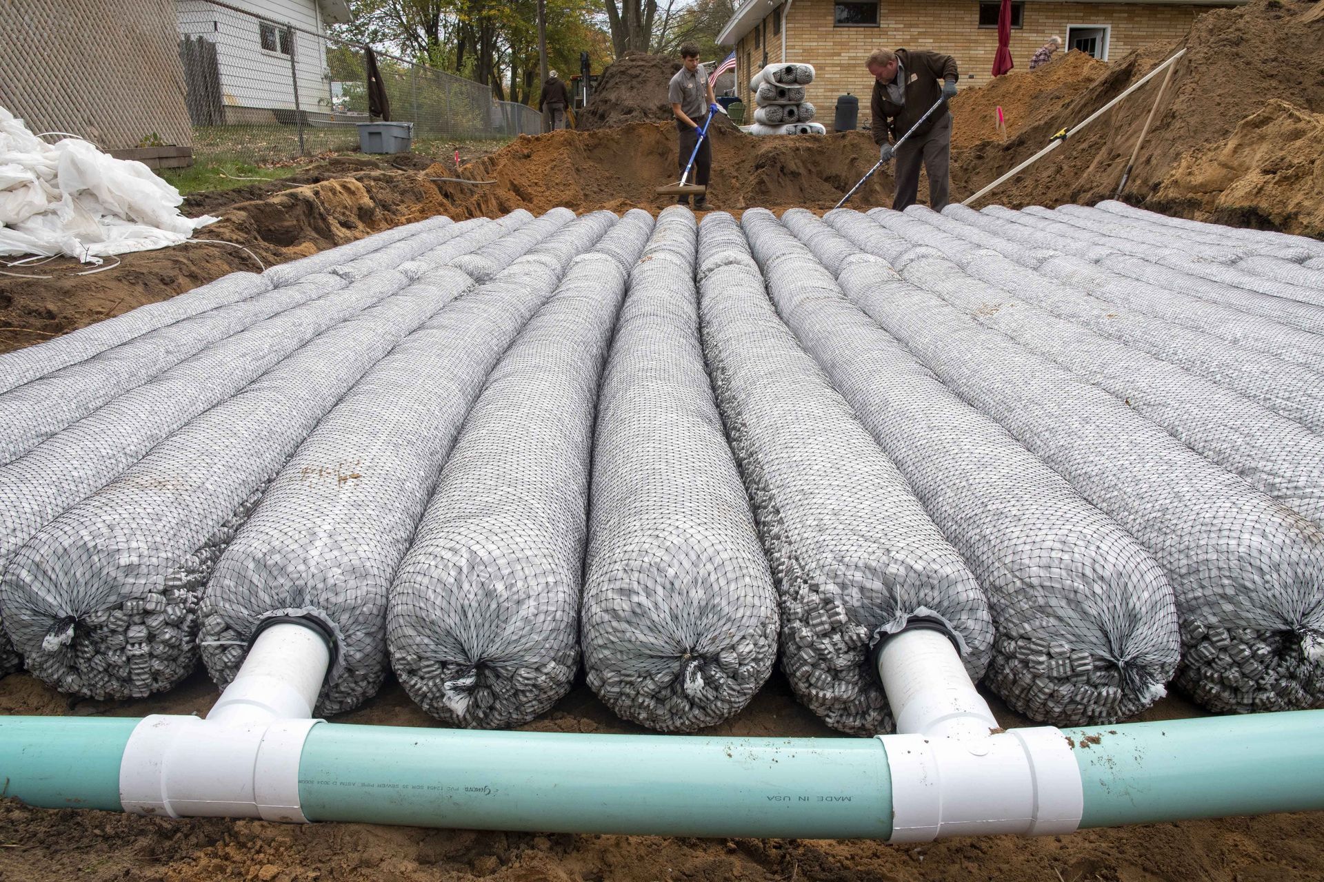 Underground septic system installation: fabric tubes filled with gravel connected to a PVC pipe in a dirt trench.