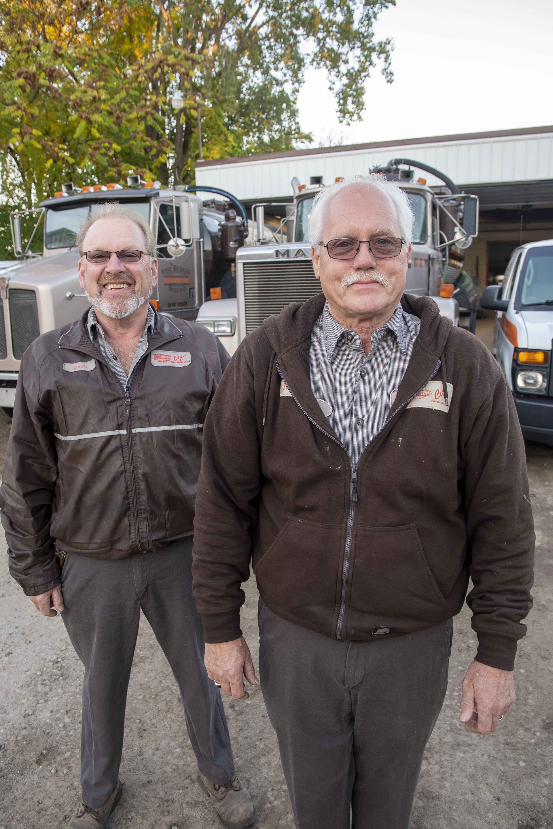 Two men standing in front of trucks. Older man in brown hoodie, younger man in brown jacket, both smiling. Outdoors.
