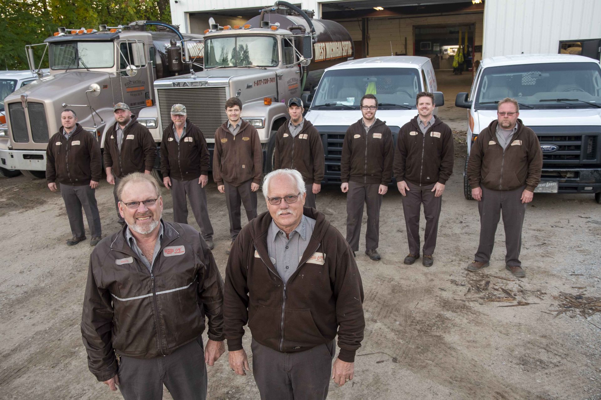 Group of workers in brown jackets pose in front of trucks and vans in a gravel lot.