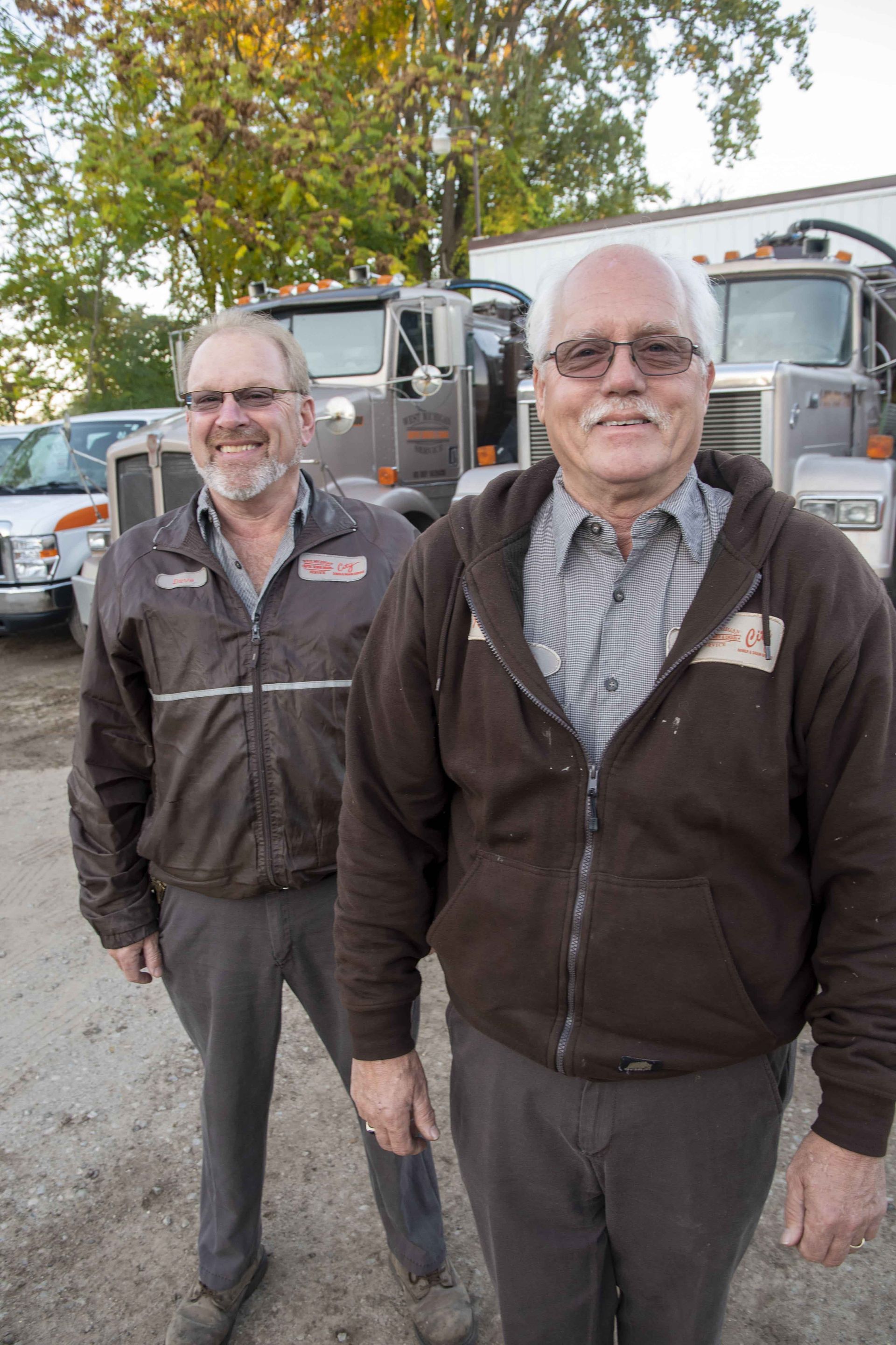 Two men in brown jackets stand near trucks, smiling outdoors.