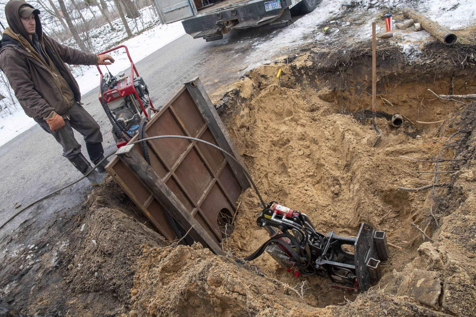 Man operating equipment in a deep trench, brown earth, metal structure, snow visible, outdoor setting.
