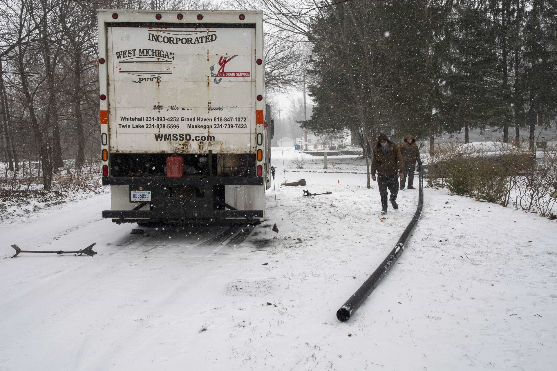 A white moving truck parked on a snow-covered road; two people carry a black hose away from the truck.