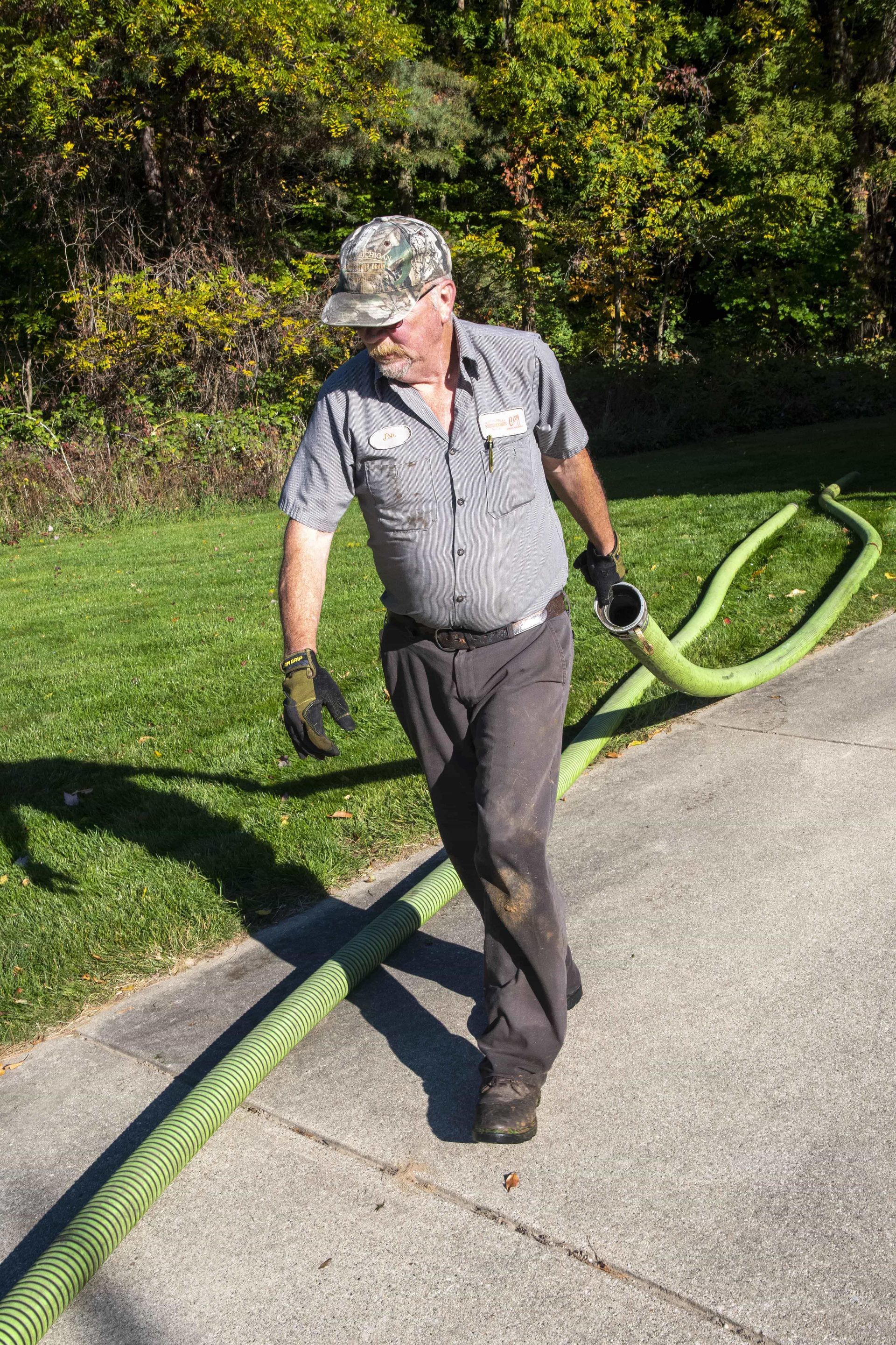 Man in uniform, holding a green hose, walking on asphalt next to grass; outdoor setting.