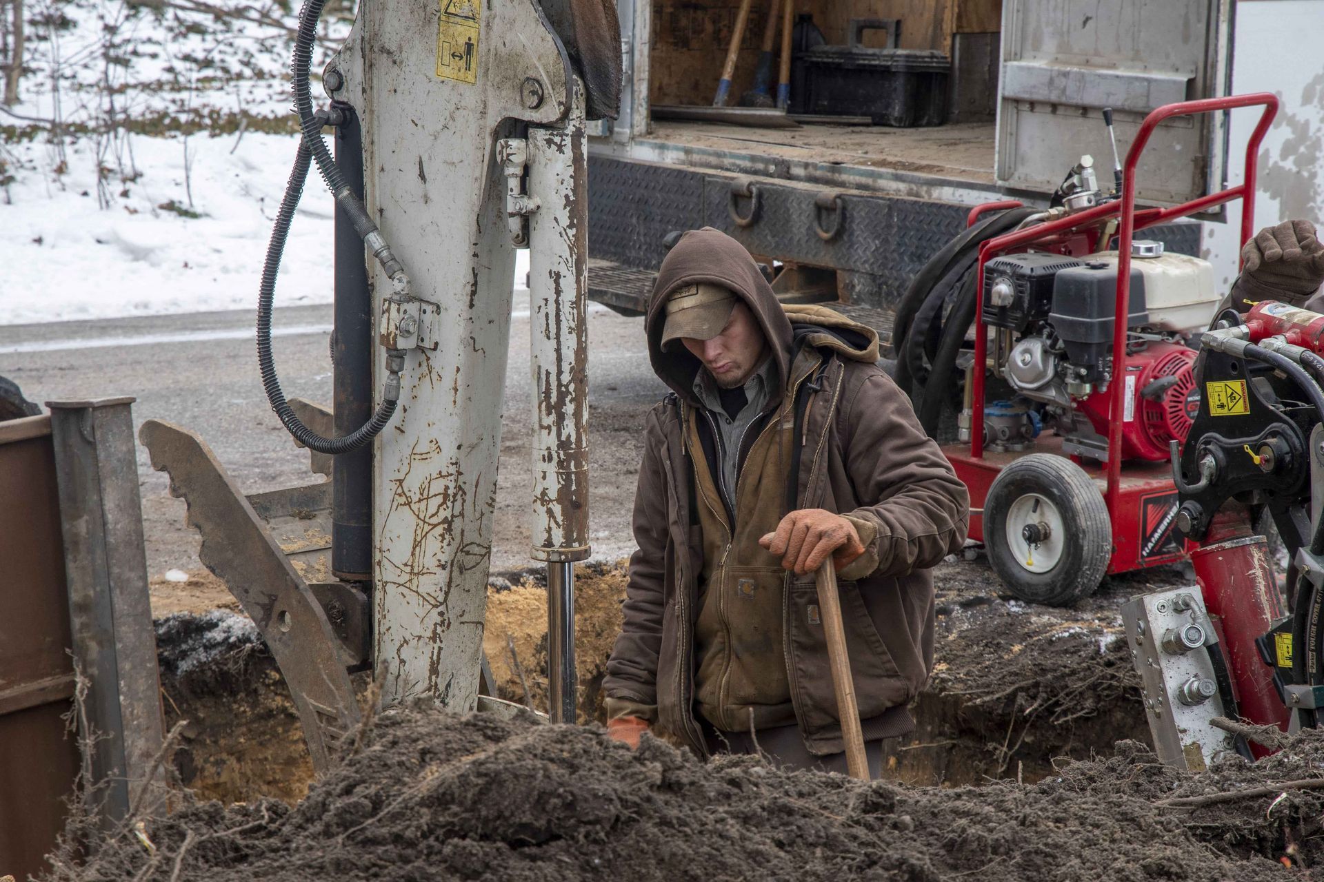 Man digging in a trench next to machinery and truck, wearing a brown hooded jacket.