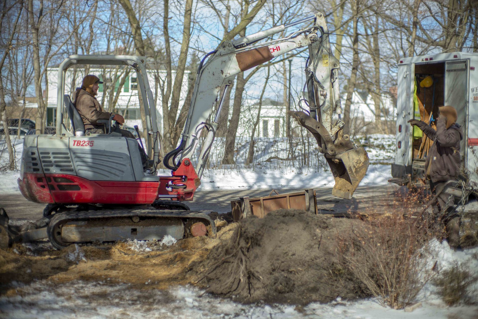 Excavator digging near a roadside with a worker nearby on a snowy day.