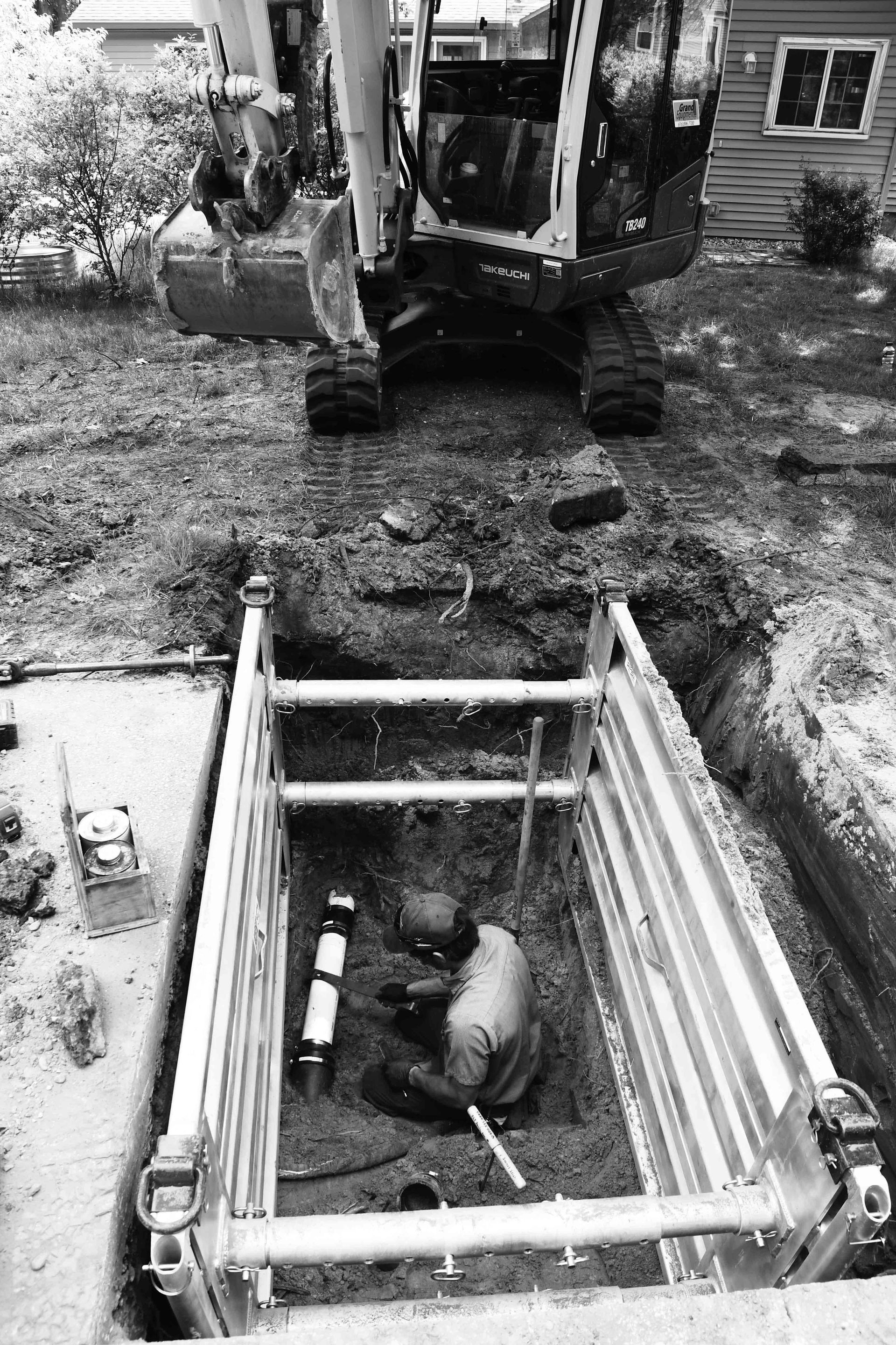 Man working in a trench, protected by a shoring system. Excavator above, grass, residential setting.
