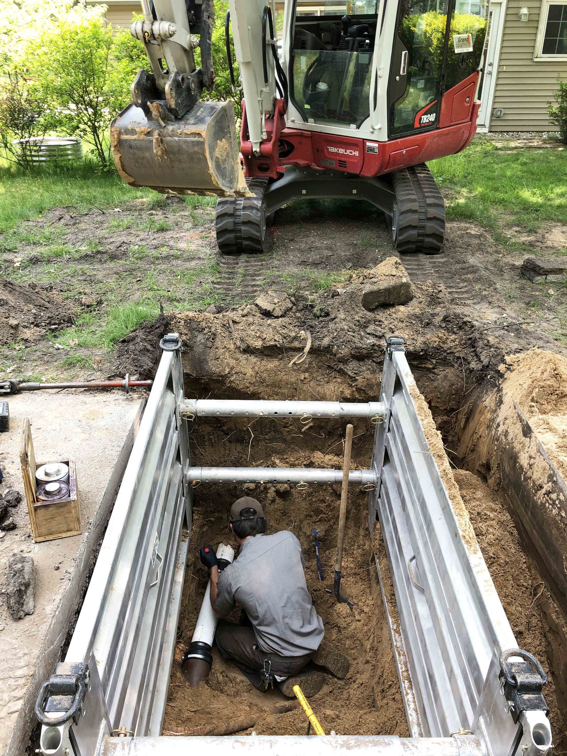 Worker in trench fixing pipe, excavator in background.