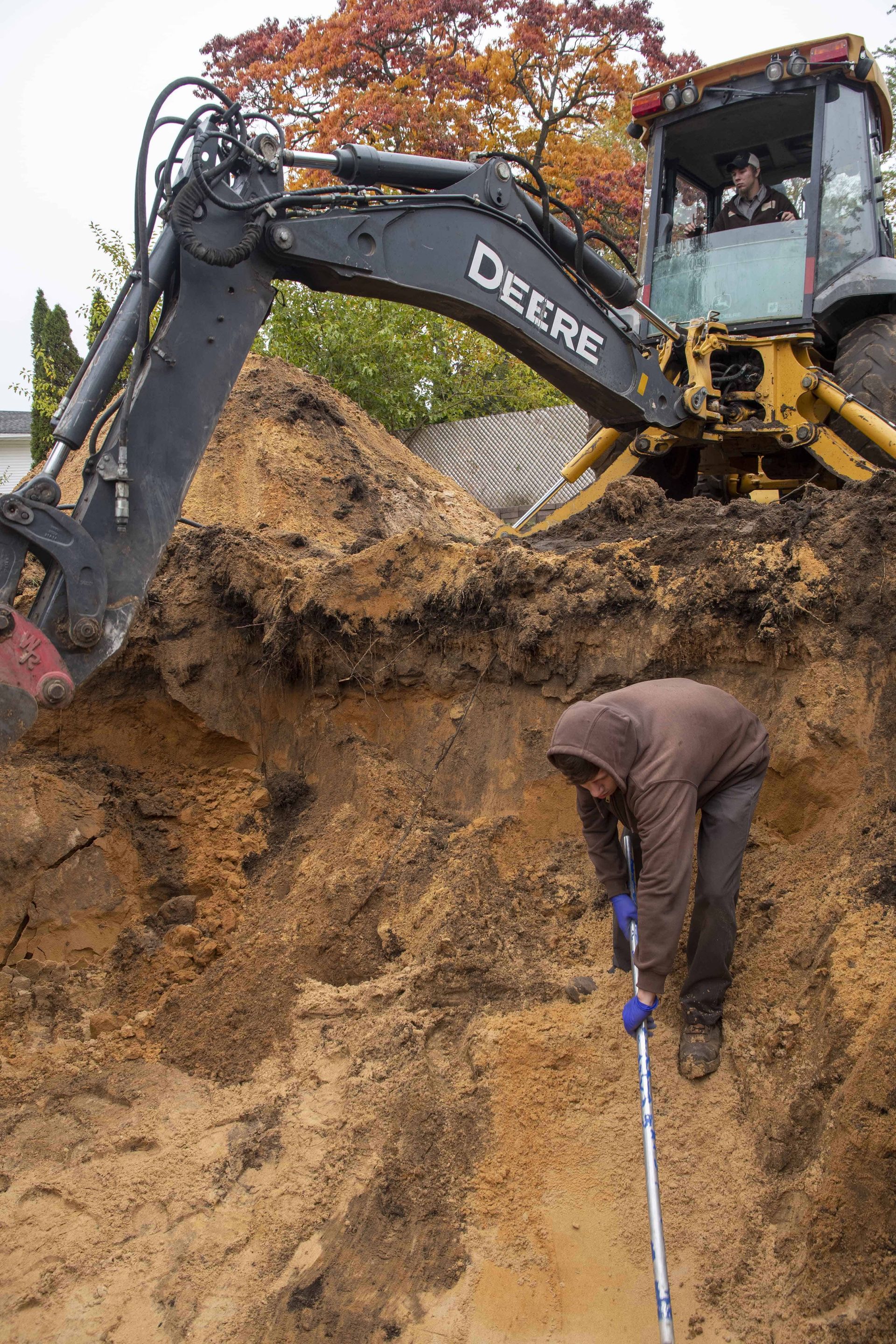 Worker digging in a trench near a John Deere excavator; autumn trees in the background.