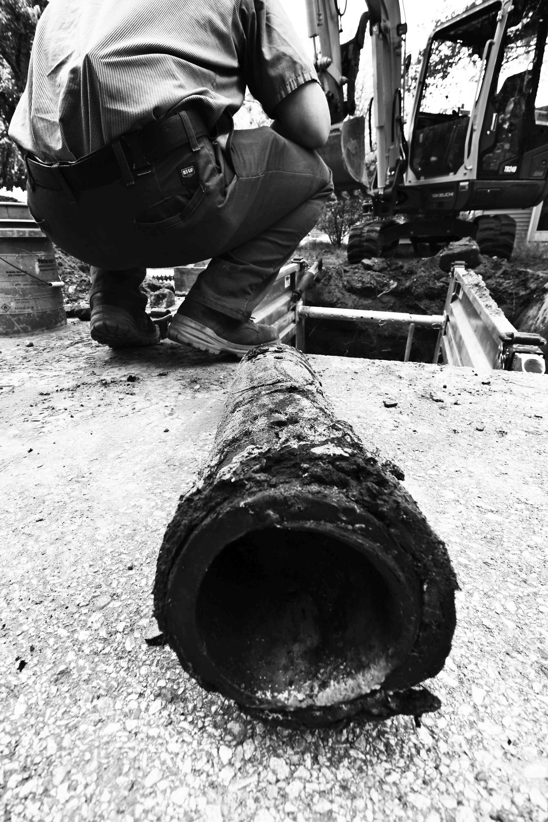 A worker inspects a corroded pipe lying in a ditch, with a backhoe in the background.