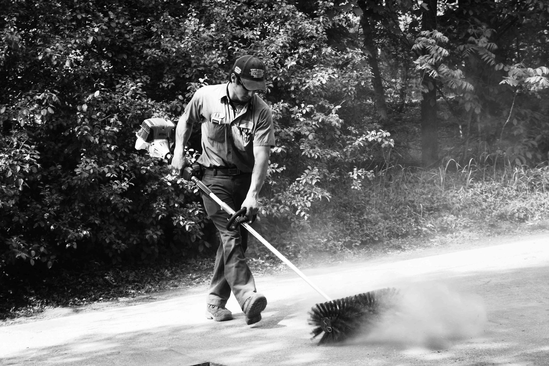 Man sweeping road with brush, creating dust cloud. Outdoors, trees in background.
