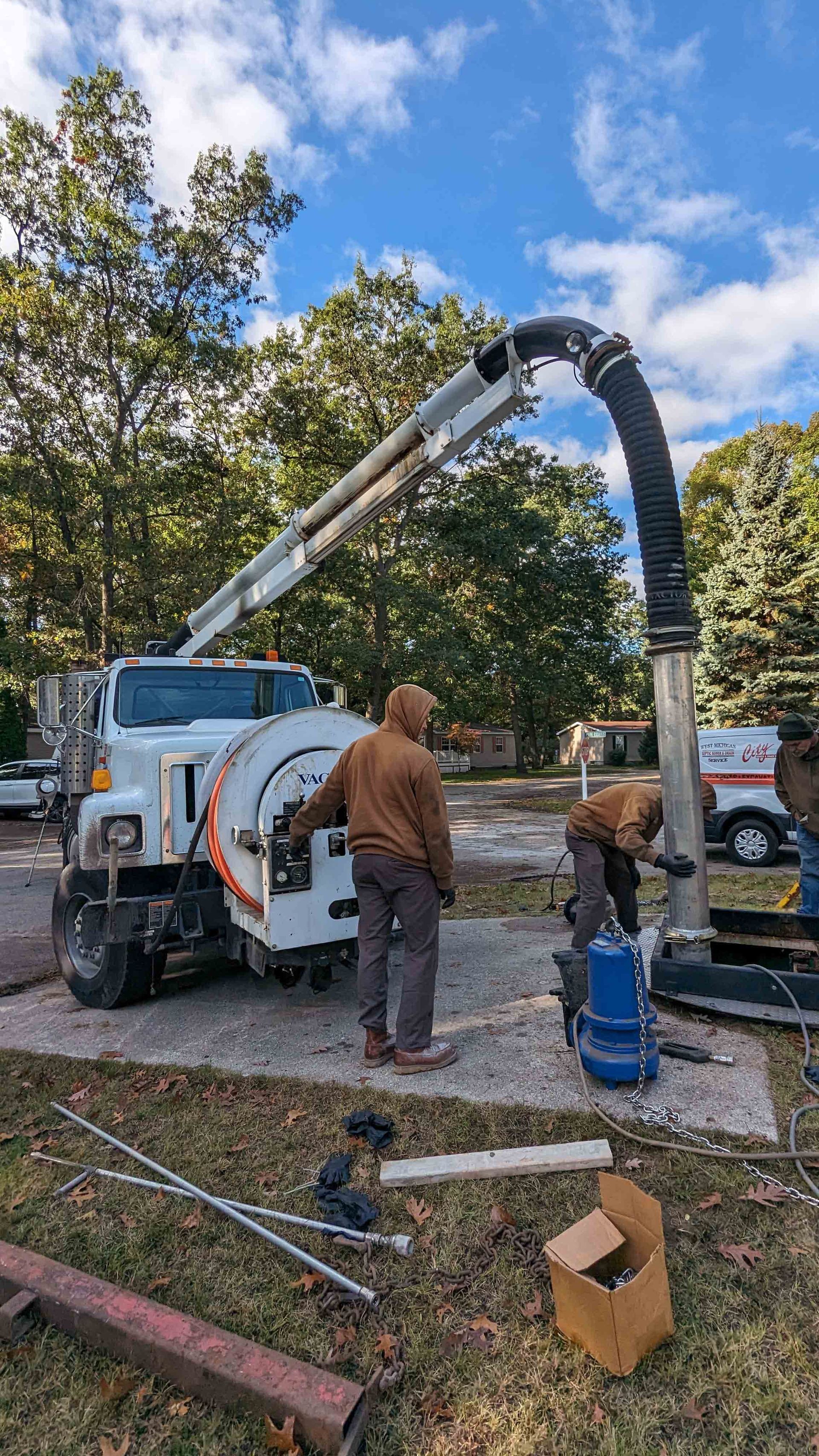 Workers using a vacuum truck to service a pump station. Blue sky and trees in background.