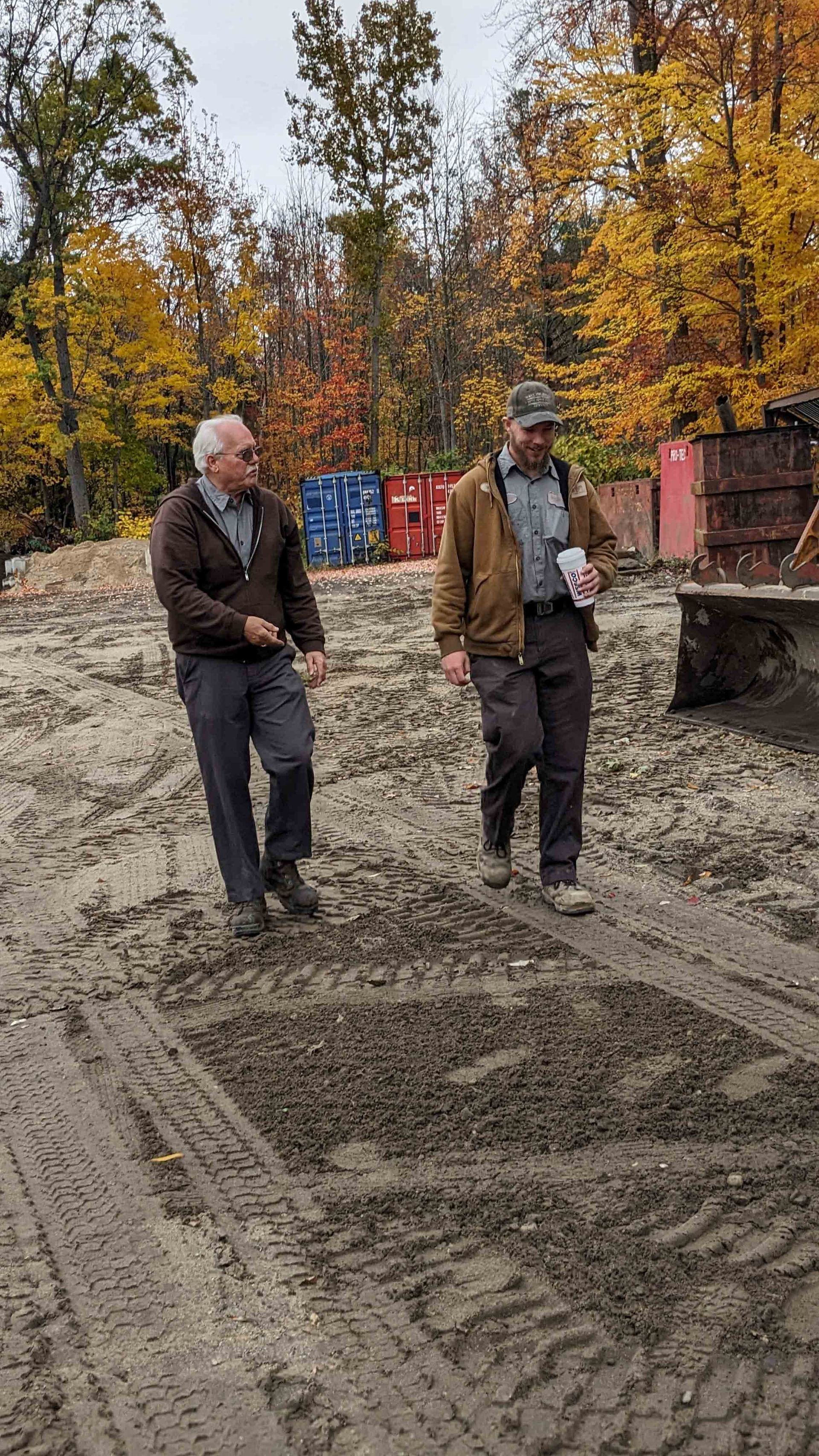 Two men walking on muddy ground, autumn trees in the background.