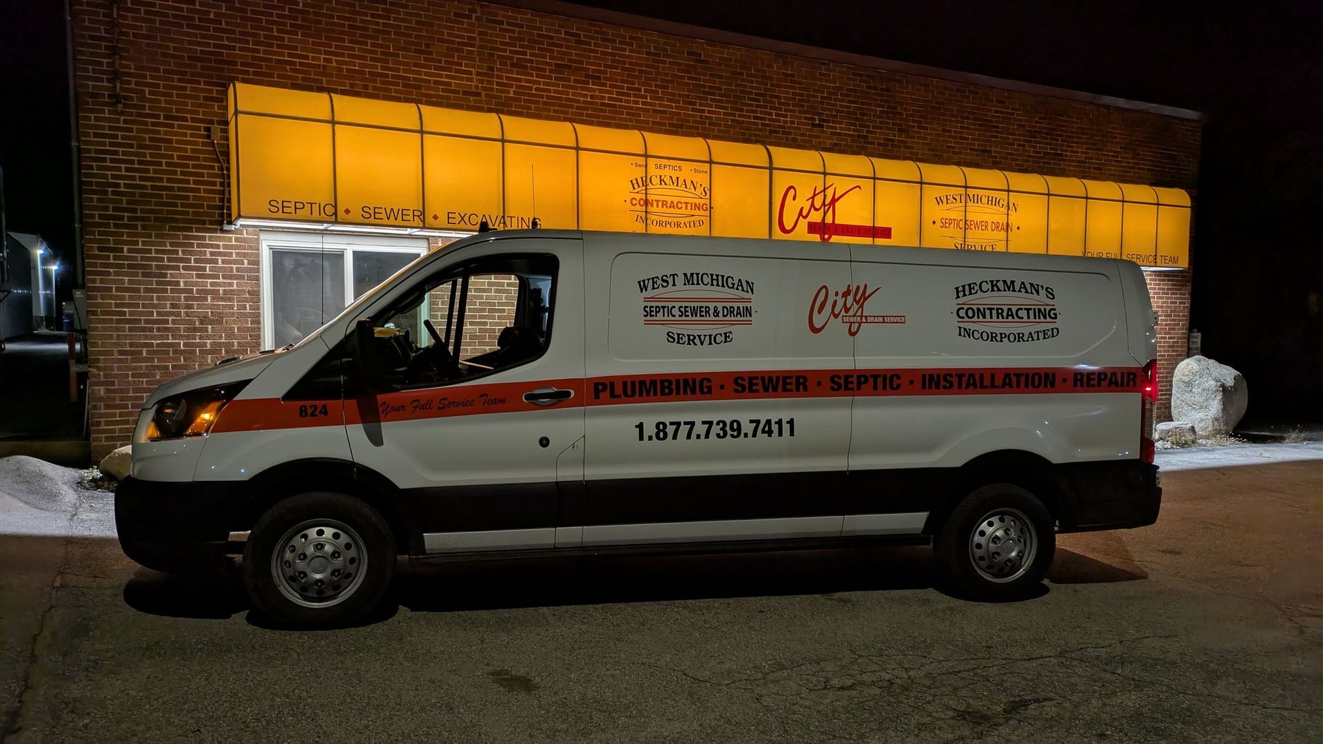 White service van with orange stripe parked in front of a brick building with yellow sign; night.