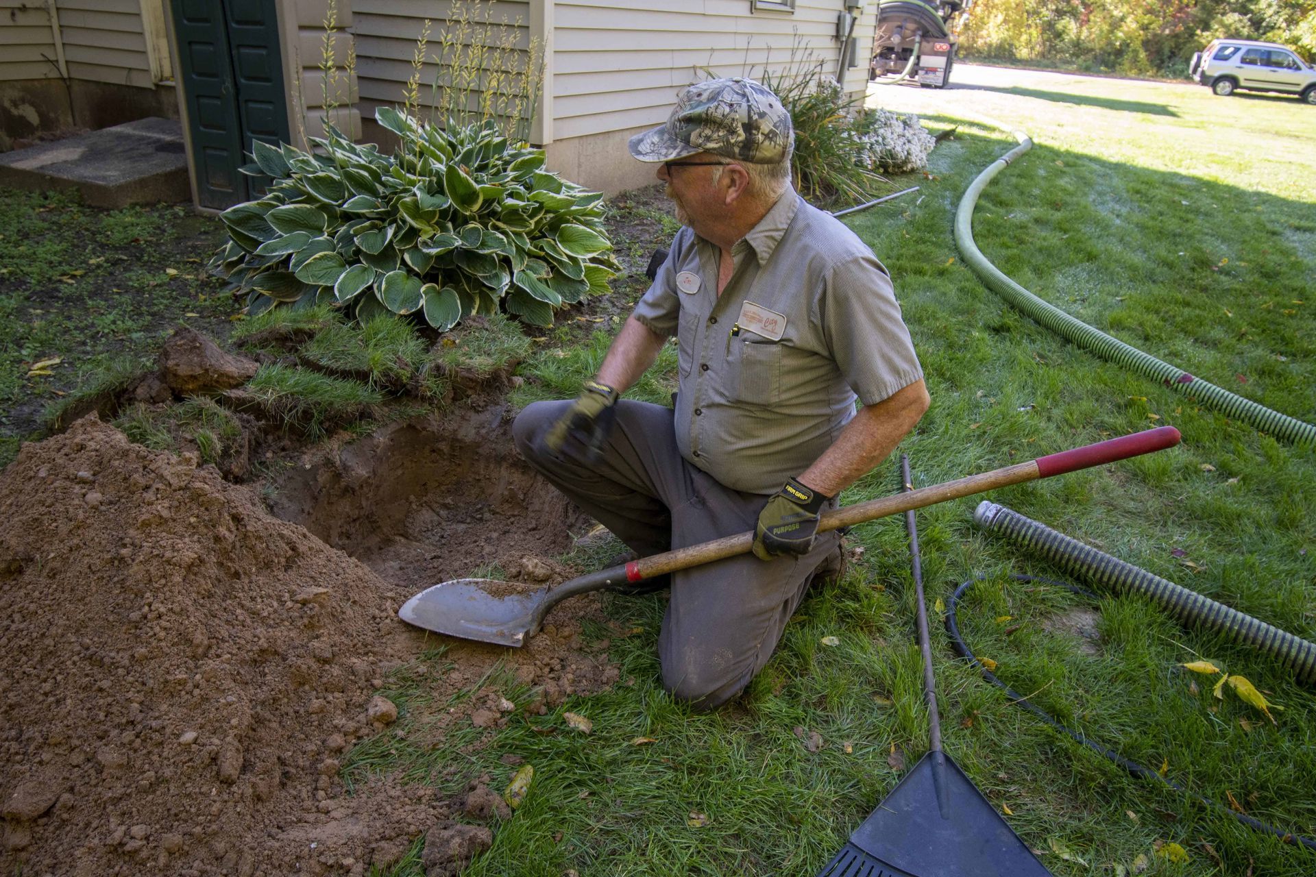 Man kneels in a hole digging with a shovel next to a house and lawn.