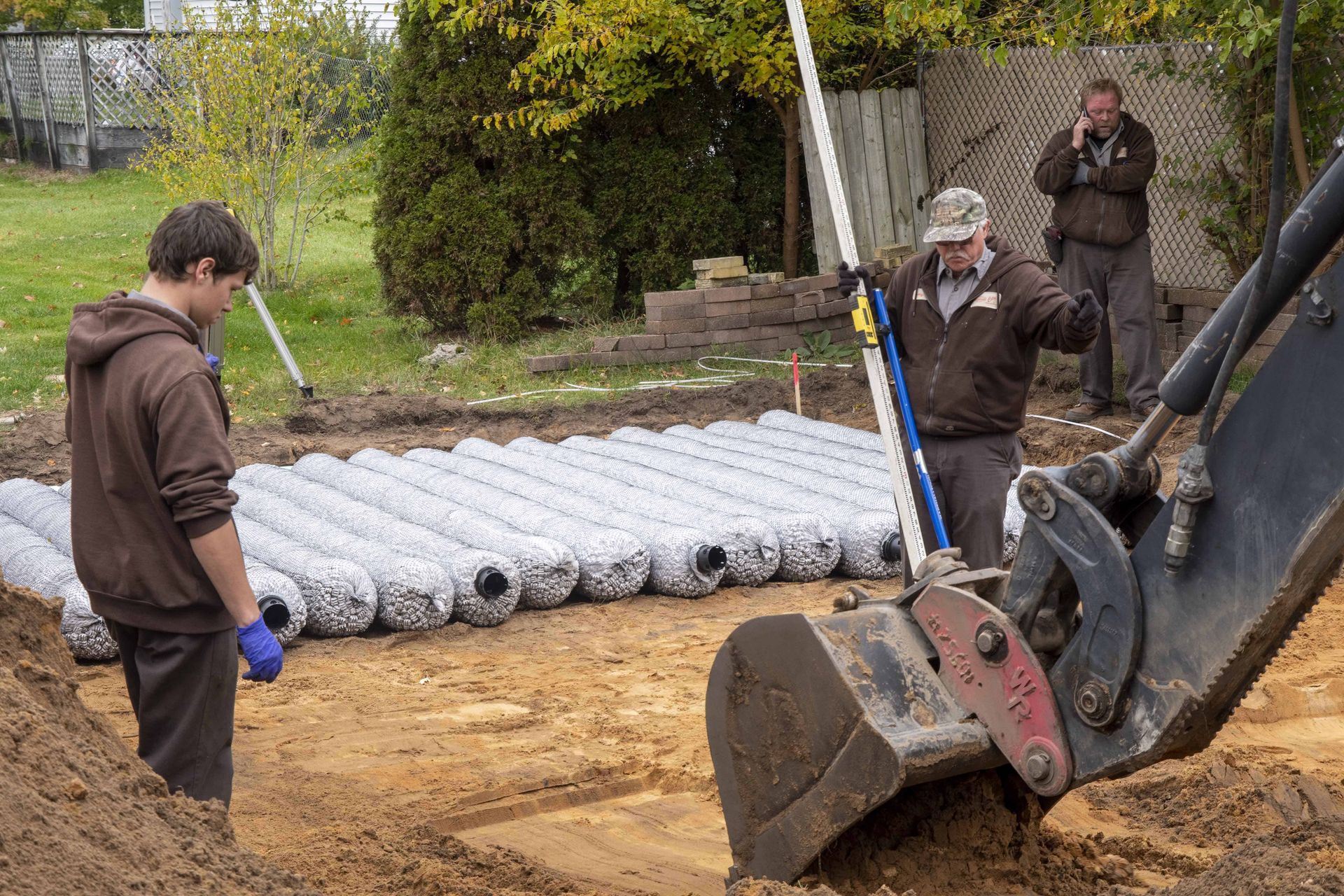 Three workers installing landscaping materials; excavator in foreground.