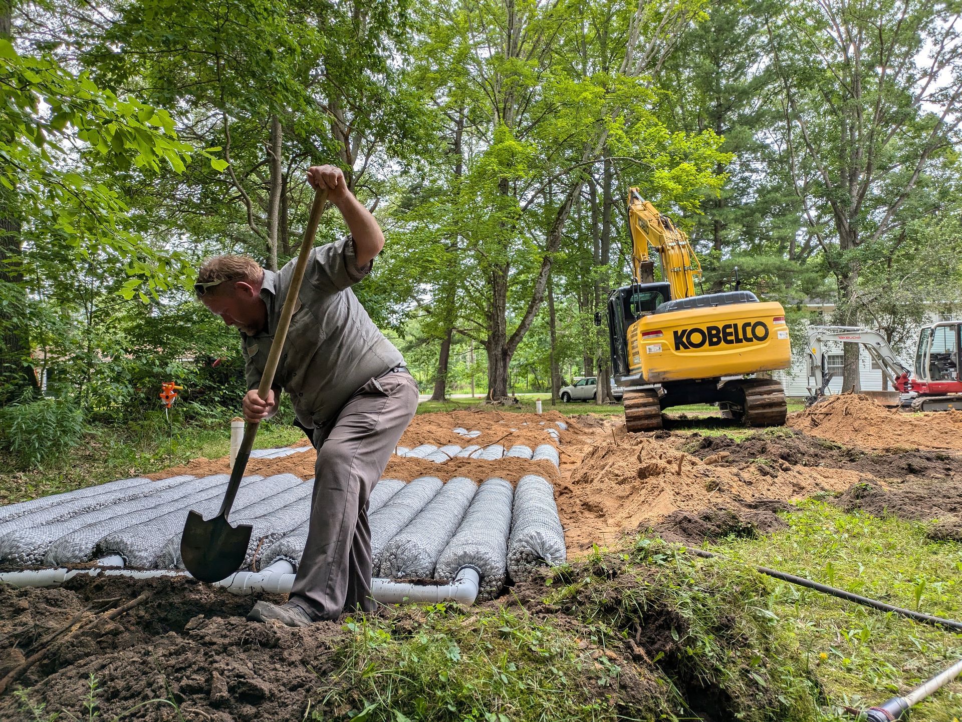 Man shovels dirt near rolled fencing, excavator in background on a construction site.