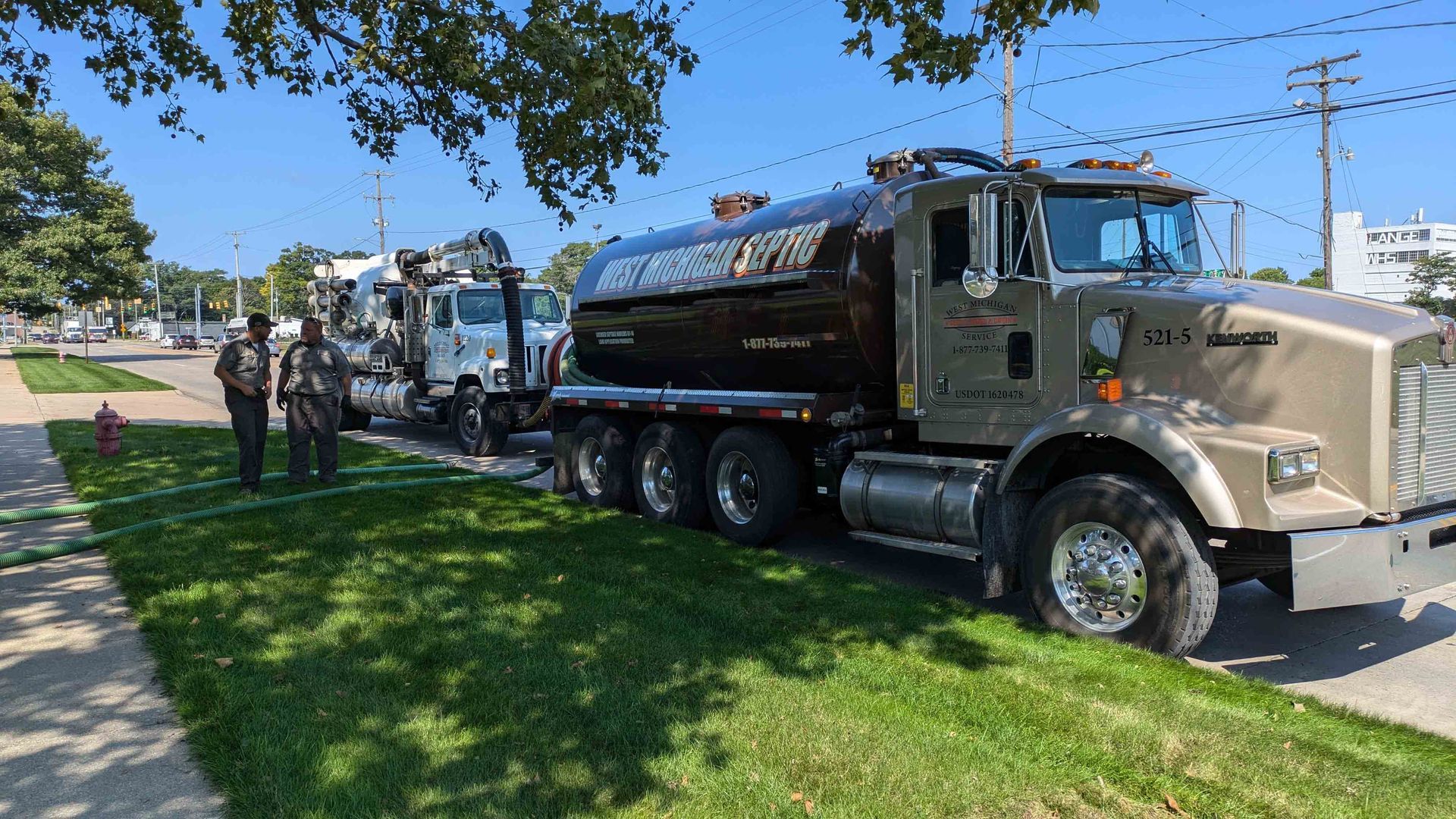 Three fuel tanker trucks parked on grass near a sidewalk, with people standing nearby.