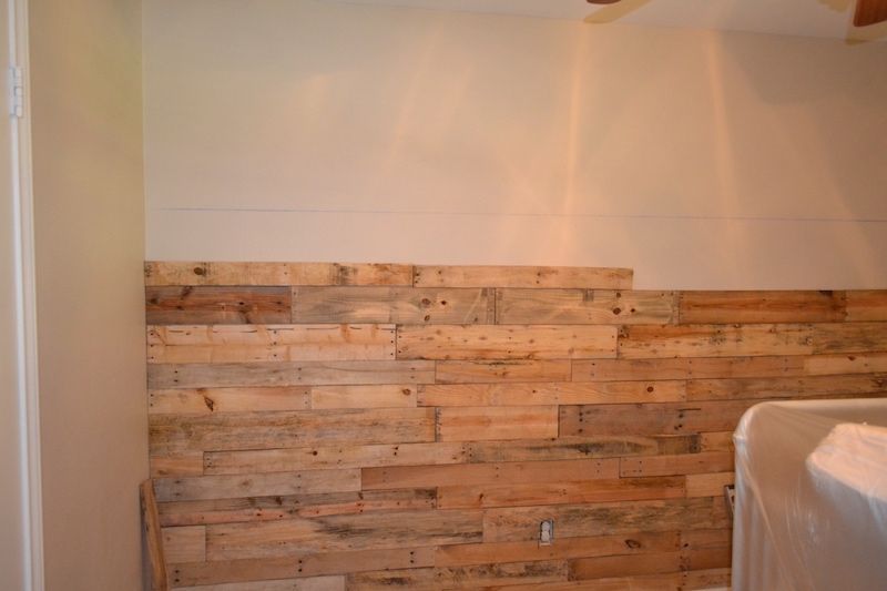 Wooden pallet wall partially installed in a room. Brown and tan wood against a white wall.