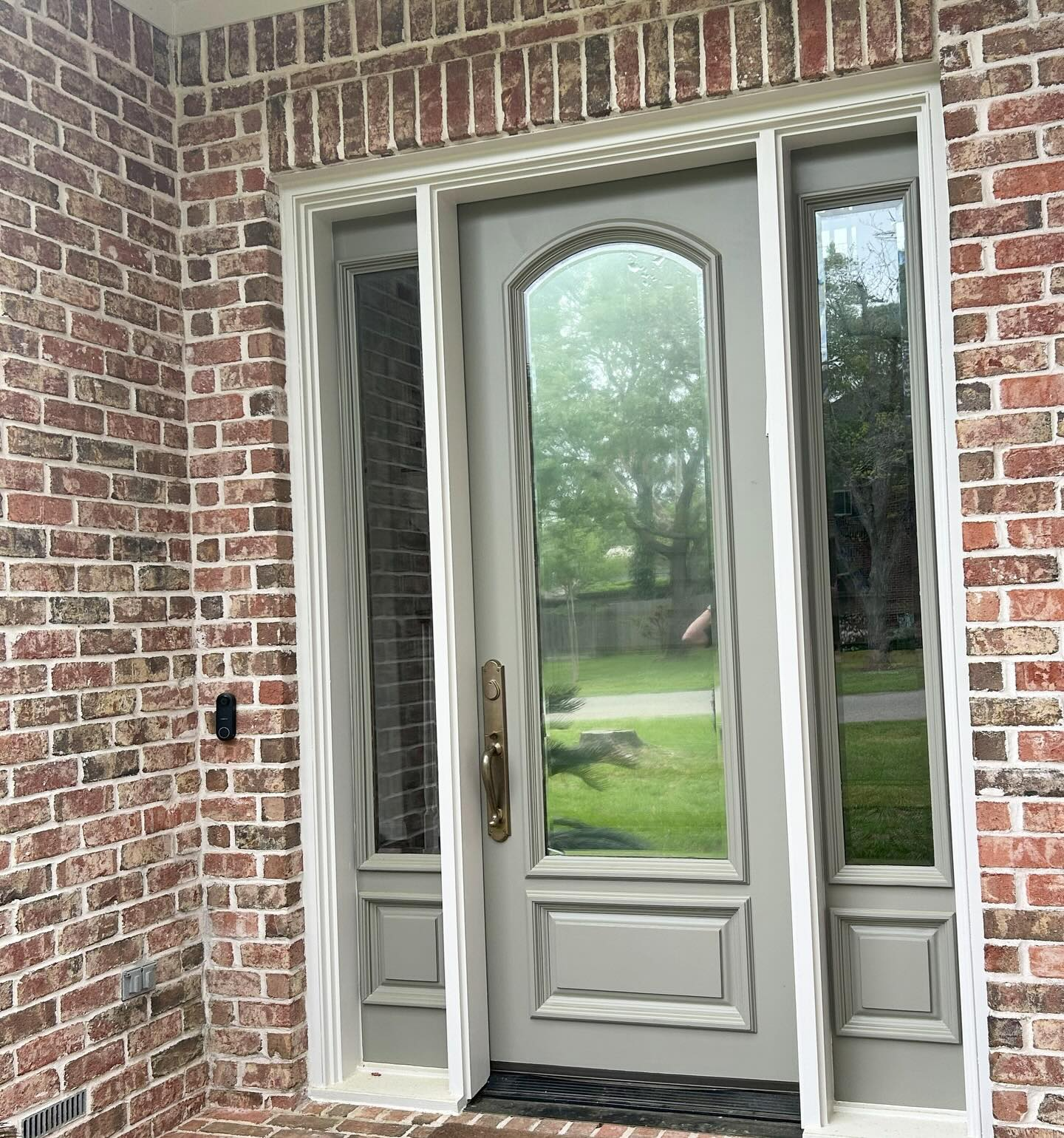 Grey front door with arched glass panel, flanked by two side windows, set in a brick exterior.