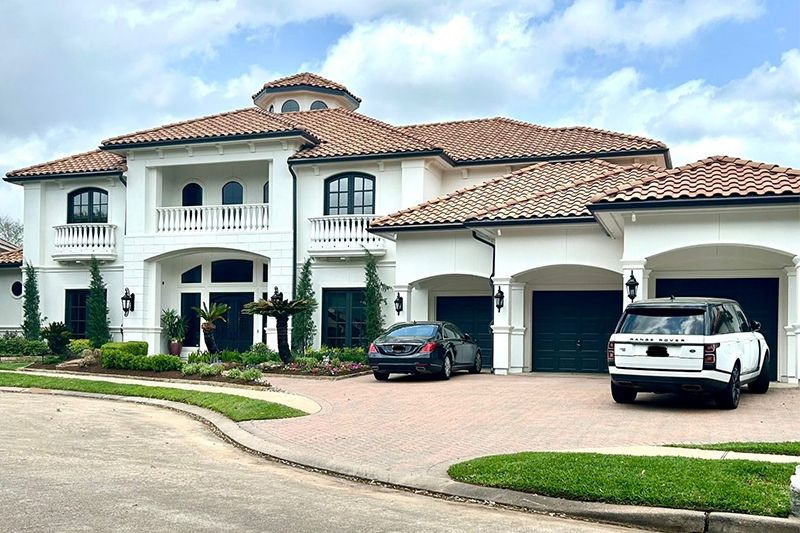 White stucco mansion with a terracotta roof, arched windows, and a circular entry. Two cars parked in the driveway.