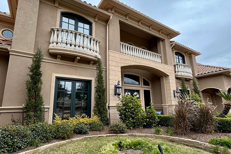 Beige two-story house with balconies, arched doorways, and dark window frames. Landscaping in front.