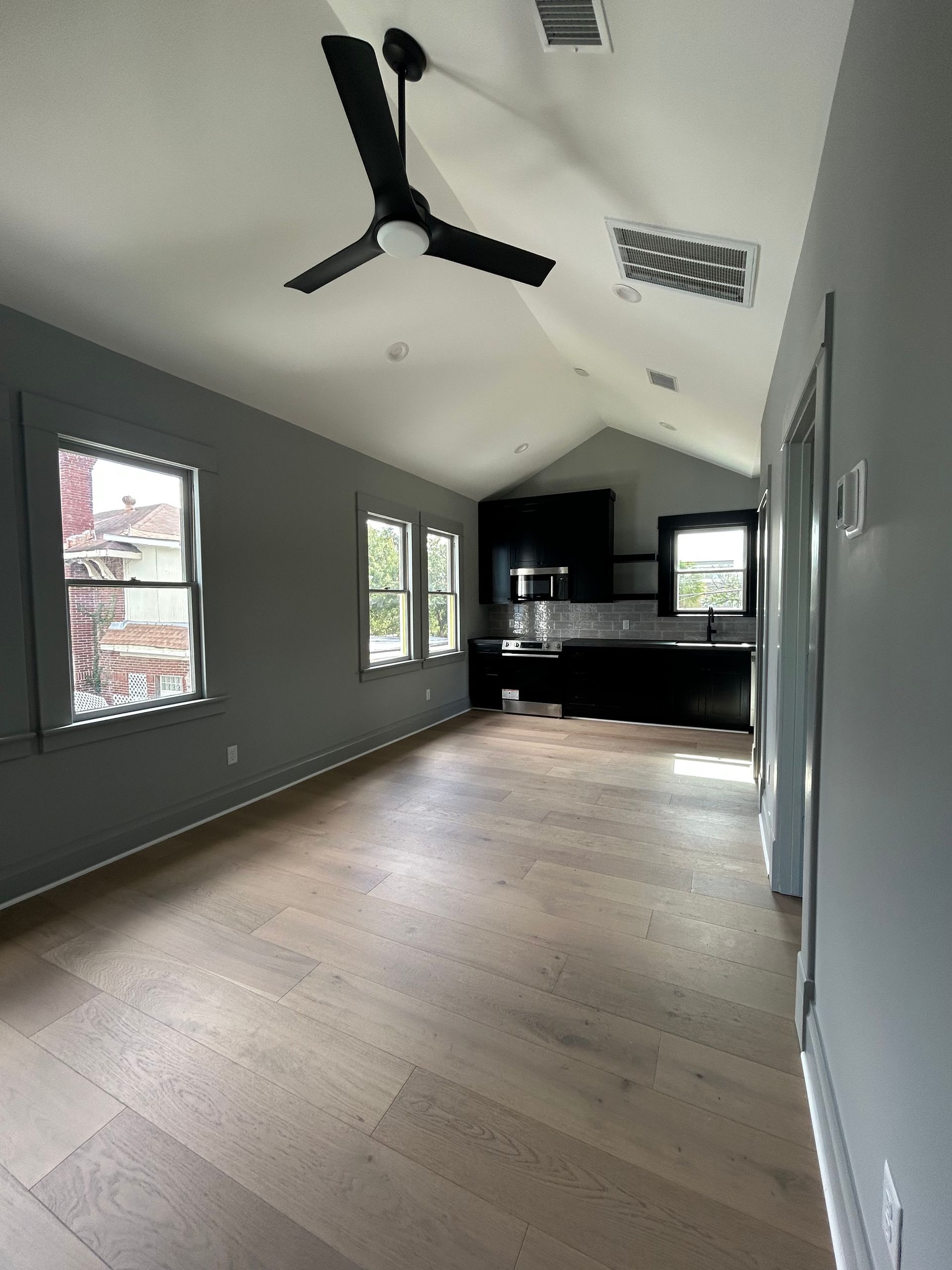 Empty modern living room with gray walls, wood floors, high ceiling, ceiling fan, and kitchen nook at the back