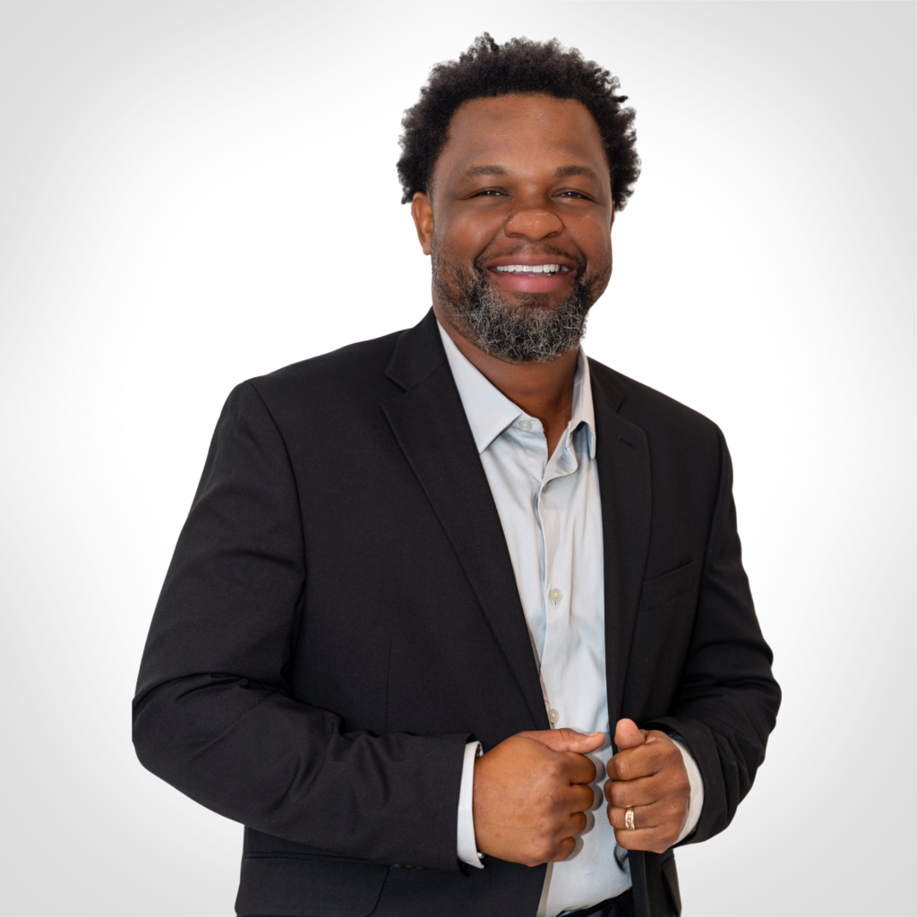Smiling man in a black suit jacket, holding the lapels against a plain white background.
