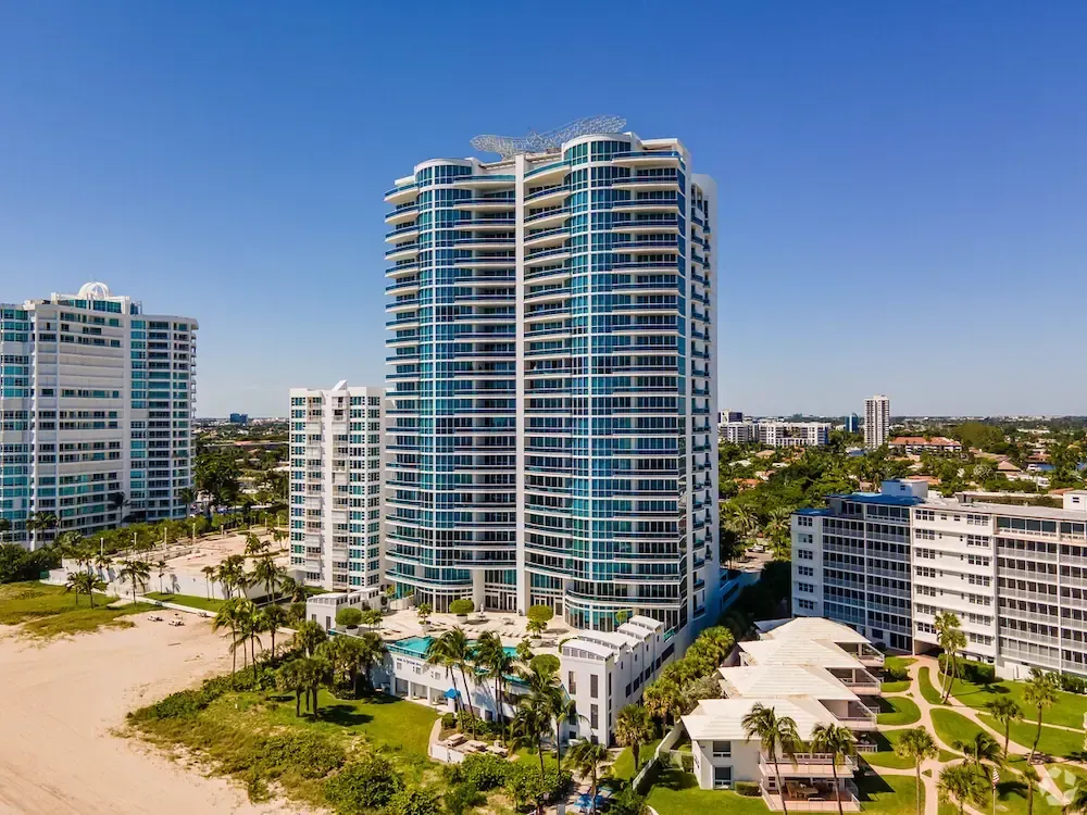 Beachfront high-rise condos beside a sandy beach and palm trees under a clear blue sky