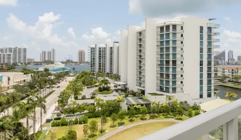 Modern waterfront apartment towers with balconies, palm-lined paths, and a canal under a bright blue sky