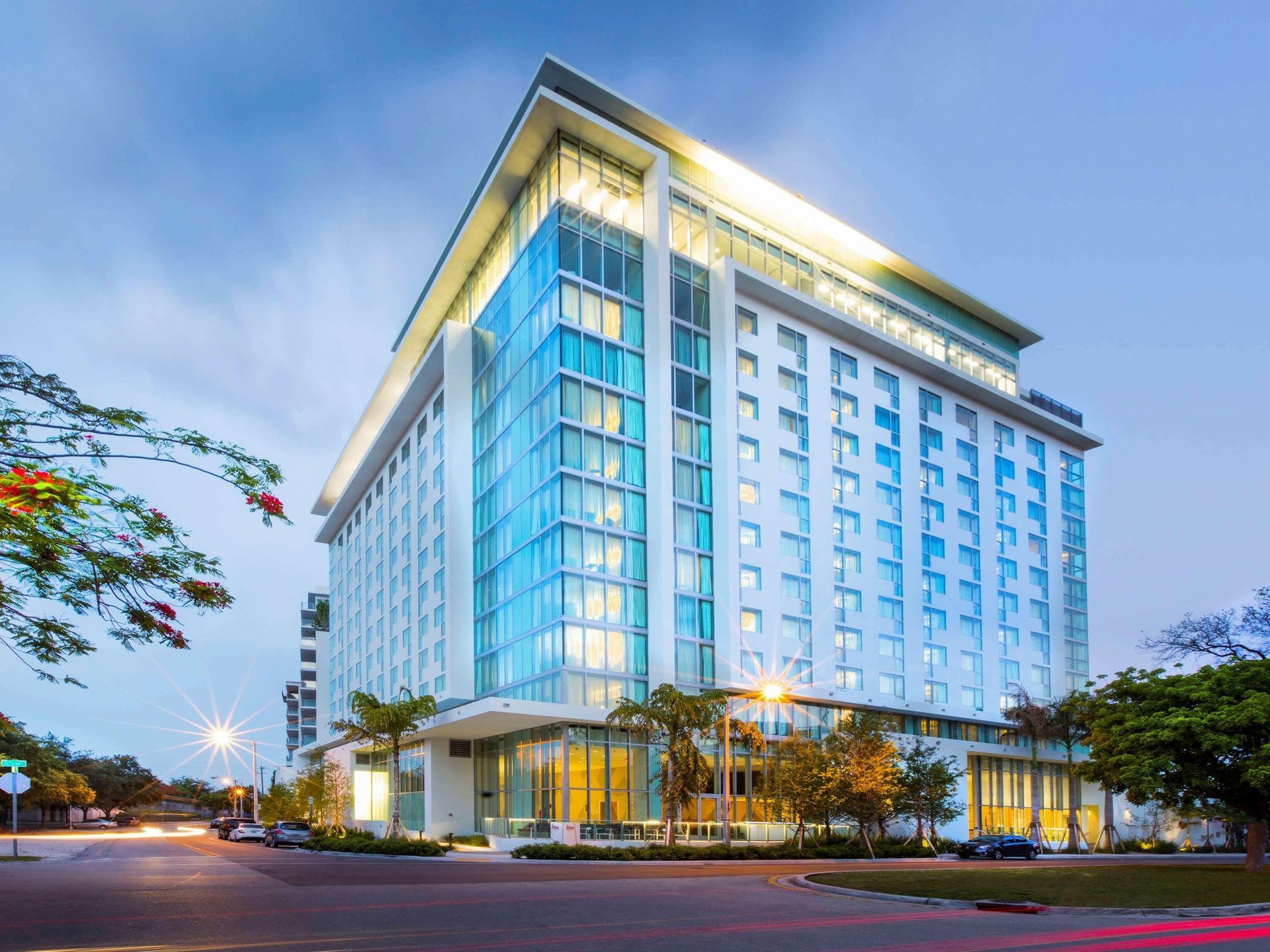 Modern glass-front hotel tower at dusk with illuminated roofline and trees in front