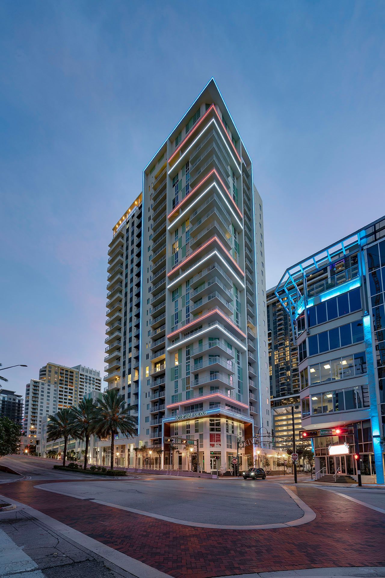 Tall modern glass office tower at dusk on a city street with palm trees and lit windows