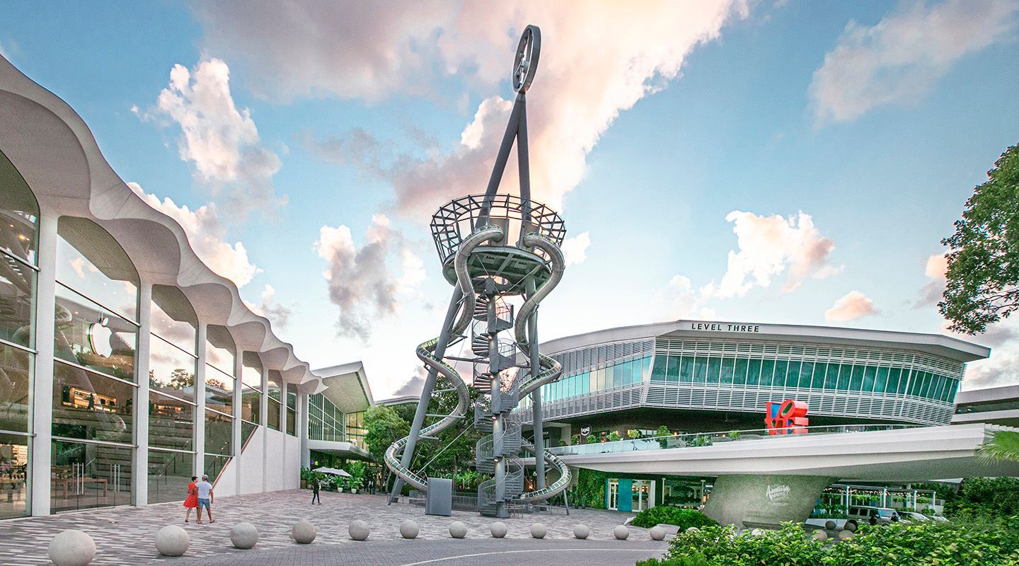 Modern sculpture in a landscaped plaza beside a curved glass building under a partly cloudy sky