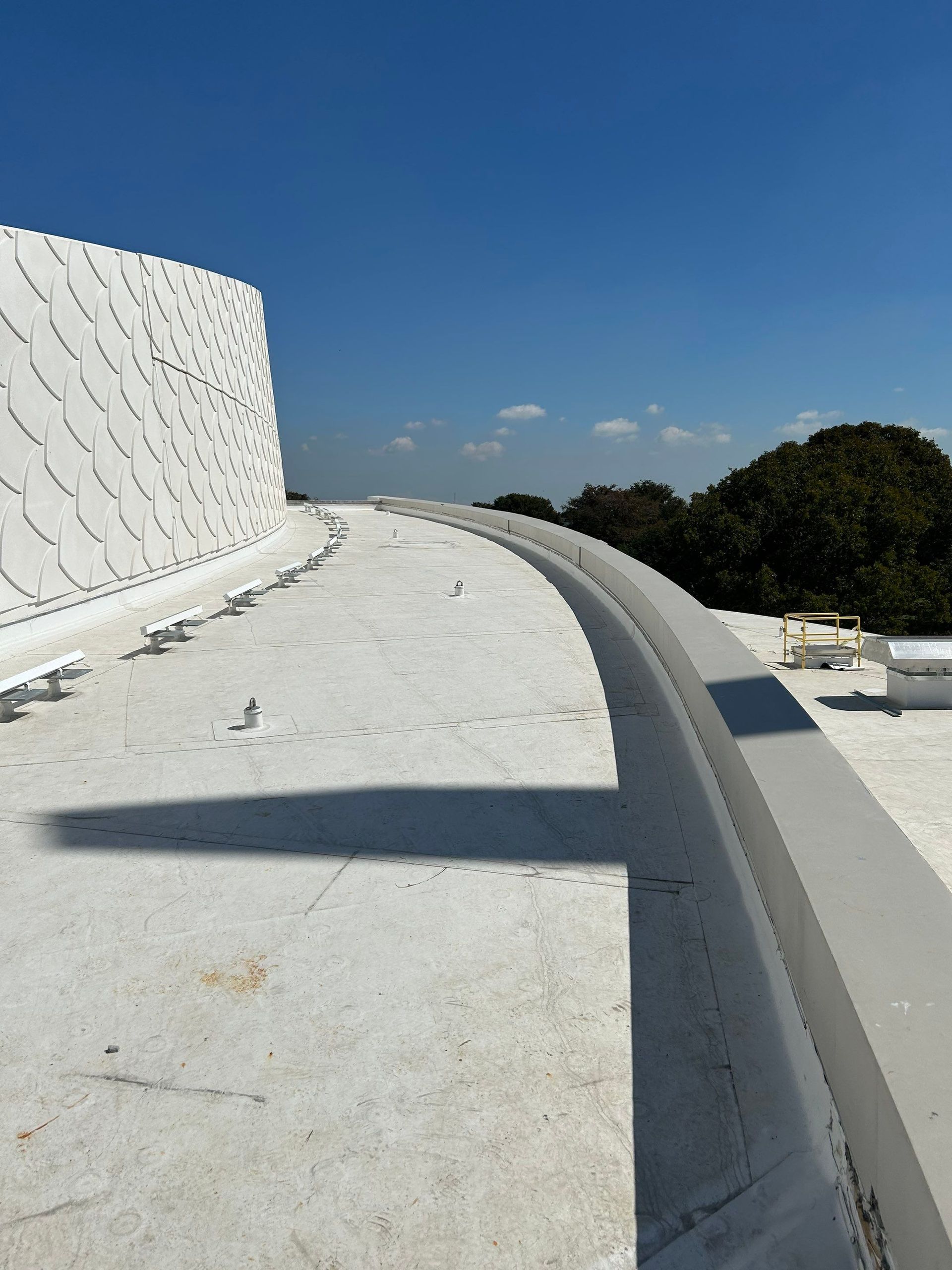 White curved rooftop walkway with a textured wall, blue sky, and trees in the distance