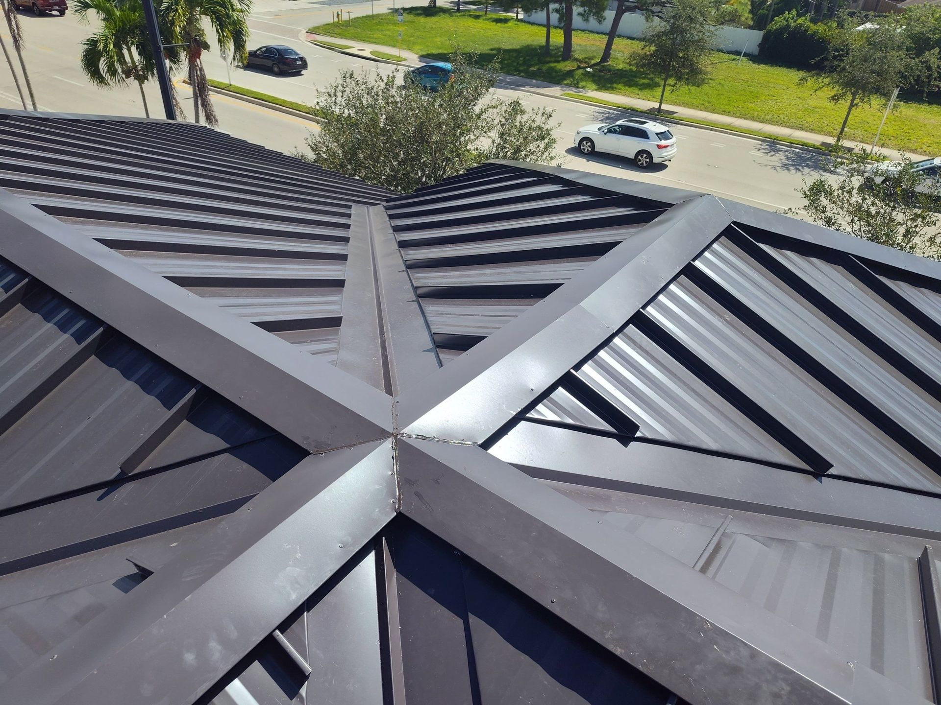 Dark metal roof with a central ridge intersection, viewed from above near a street and parked cars.