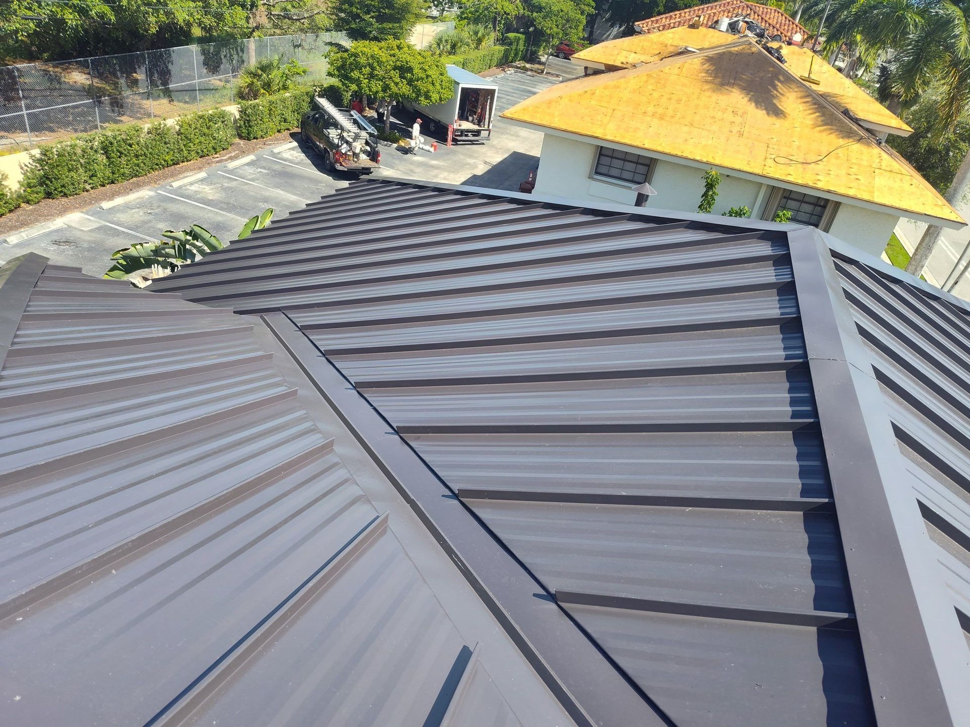 Gray metal roof panels on a house, with a yellow-roofed building and trees in the background