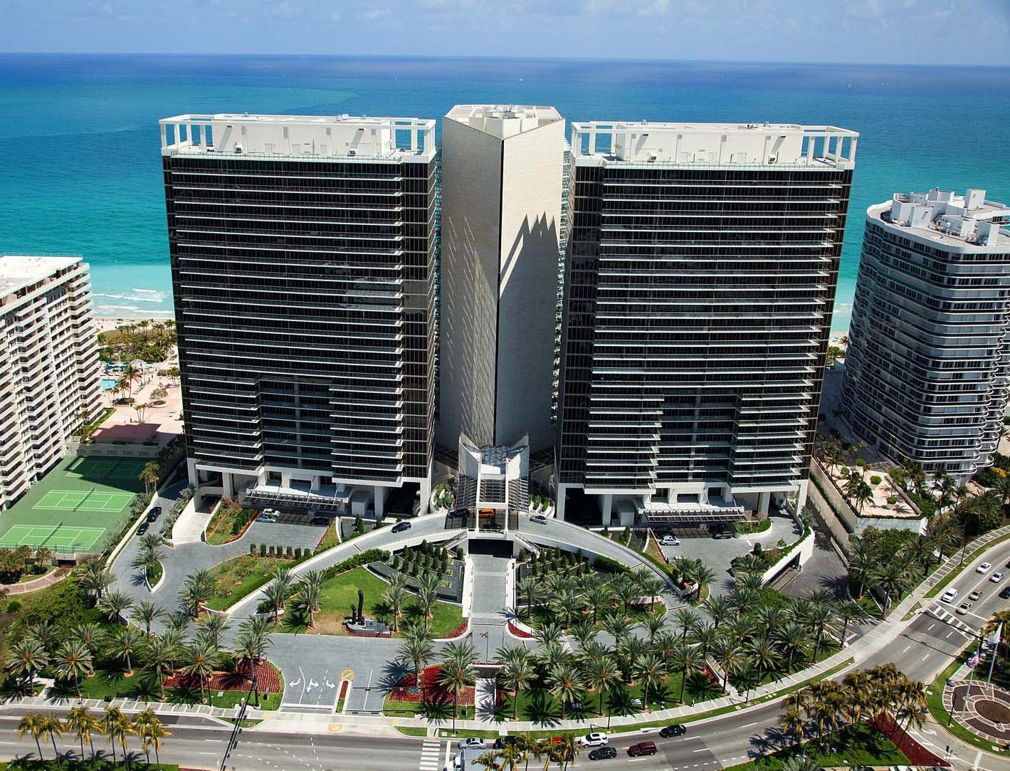 Aerial view of two modern beachfront high-rises beside turquoise ocean and landscaped grounds.