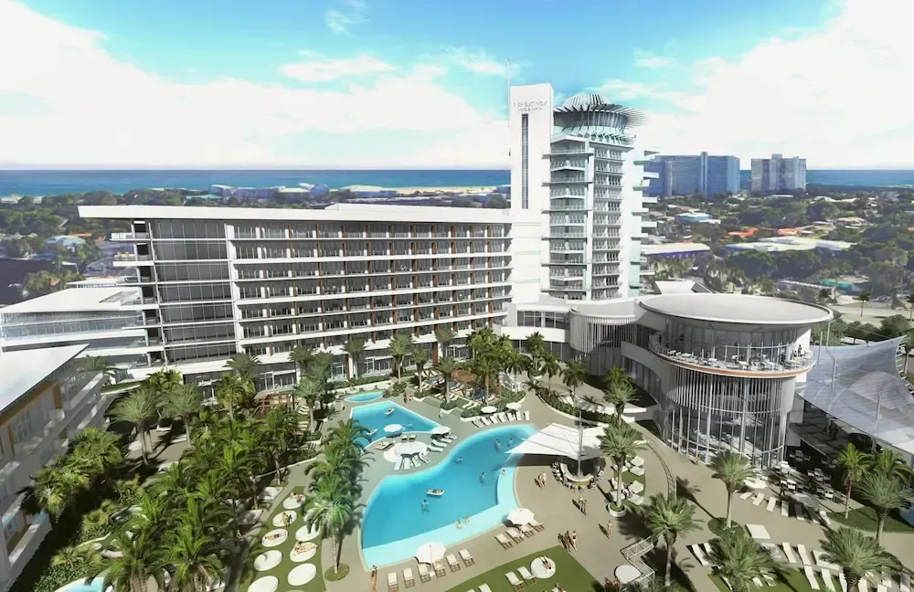 Aerial view of a modern resort hotel with a pool, palm trees, and ocean in the distance