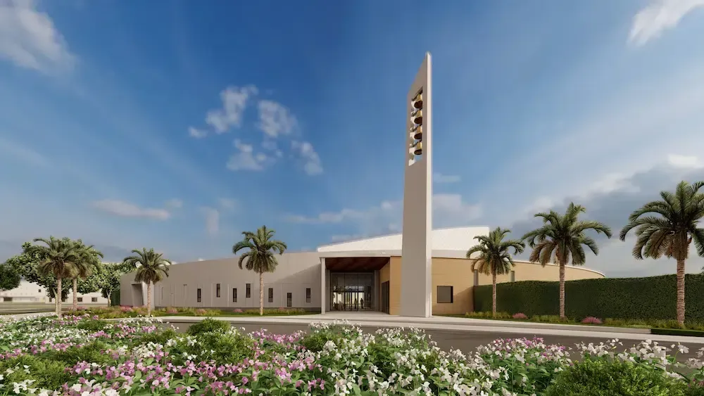 Modern church building with a tall cross tower, palm trees, and a landscaped flowerbed under a blue sky
